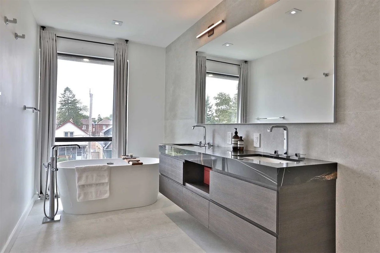 Modern bathroom featuring a white bathtub with a towel hanging over it, double sinks with a dark gray vanity, large mirror, and a window with white curtains showing trees outside.