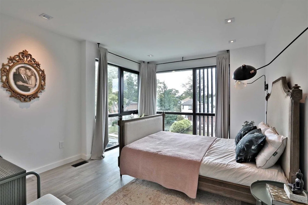 A modern bedroom with large windows, a wooden bed with beige headboard, pink blanket, black and white pillows, and a wall-mounted black reading lamp.