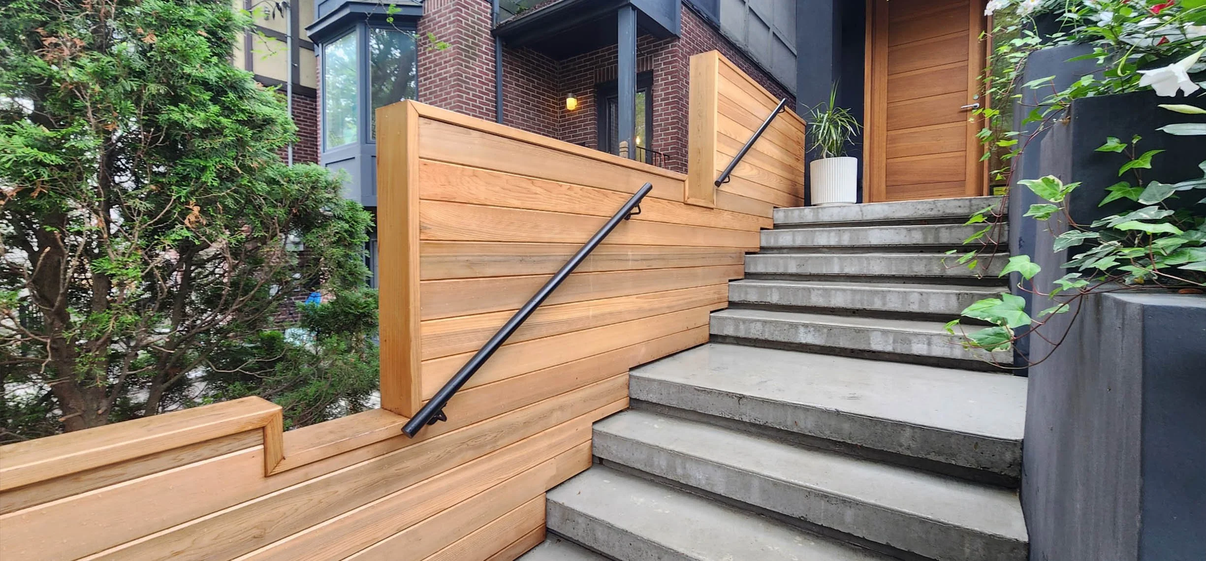 Lantern House entrance in The Beaches, Toronto, built by Maxamin Homes, featuring concrete steps, wooden door, and potted plants by flowering bushes.