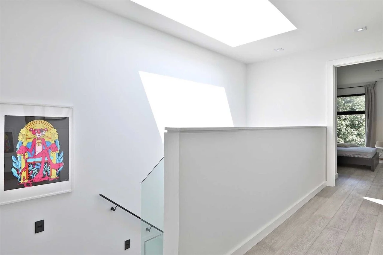 Bright, minimalist upstairs hallway with white walls, small picture of a pink flamingo in a colorful decorative frame, a large skylight, and a view into a bedroom with a large window and curtains.