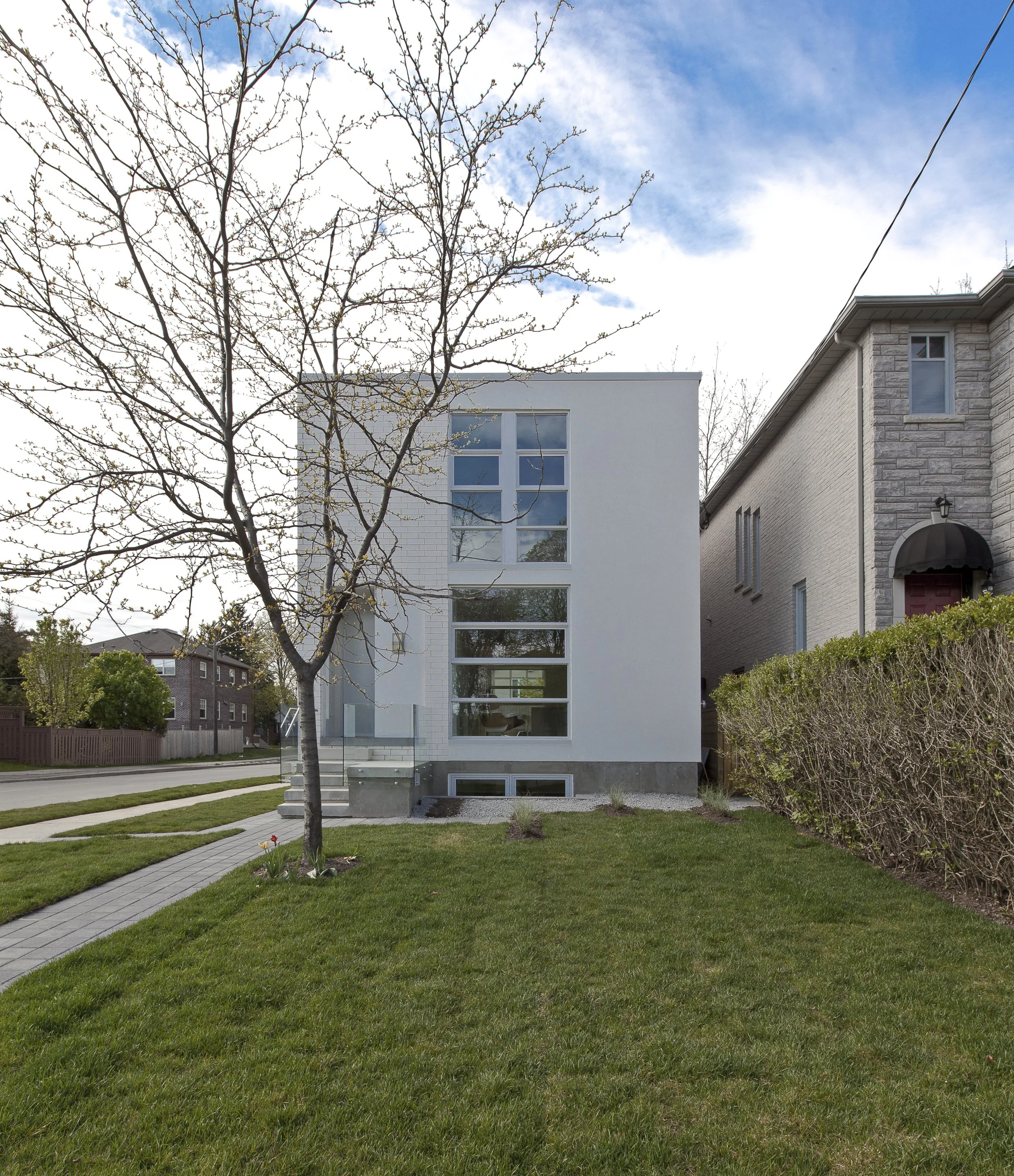 Patio House exterior in North York, Toronto, built by Maxamin Homes, modern white home with large windows and tree in front yard between brick houses.