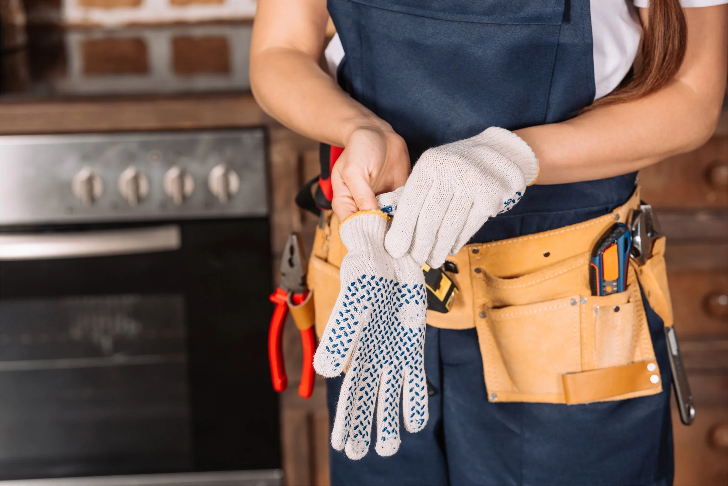 A person wearing a tool belt and gloves, holding a small object near their waist in a kitchen with an oven in the background.