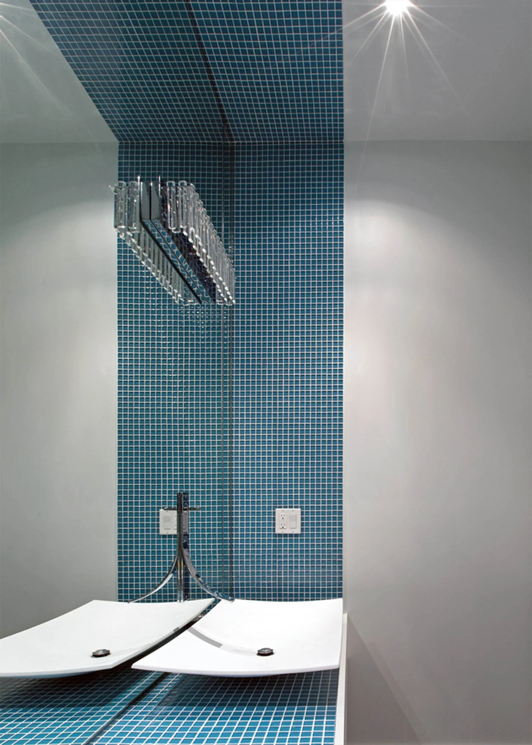 Modern medical room with blue mosaic tiled wall, two white lounge chairs, wall-mounted metal trays, and a wall outlet.