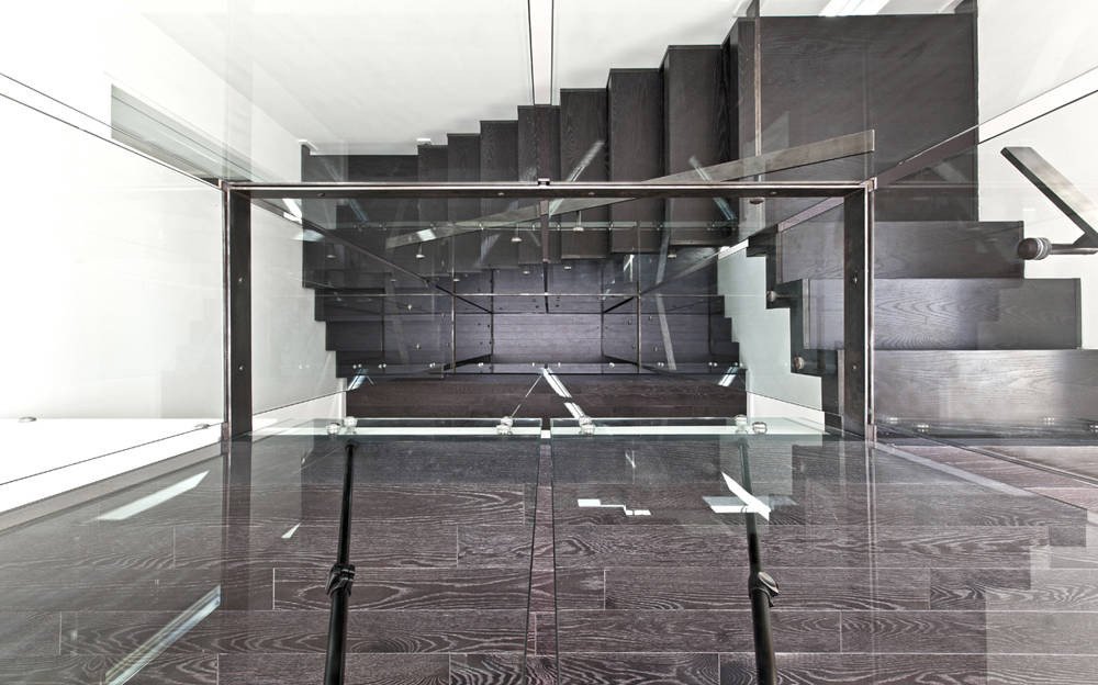 Top view of a modern staircase with dark wooden steps, glass railing, and metal handrail inside a contemporary building.