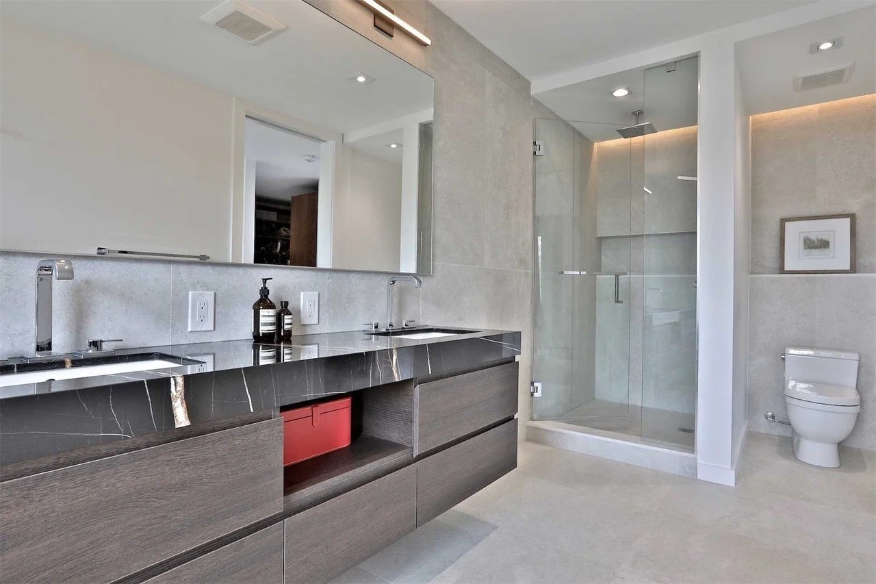 Modern bathroom with dual sinks, large mirror, shower with glass door, and toilet. Light-colored walls and flooring.