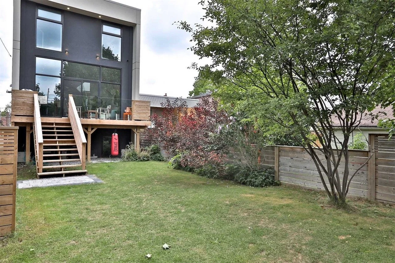 A backyard with a grassy lawn, a wooden deck with stairs, patio furniture, and various trees and bushes surrounded by a wooden fence.