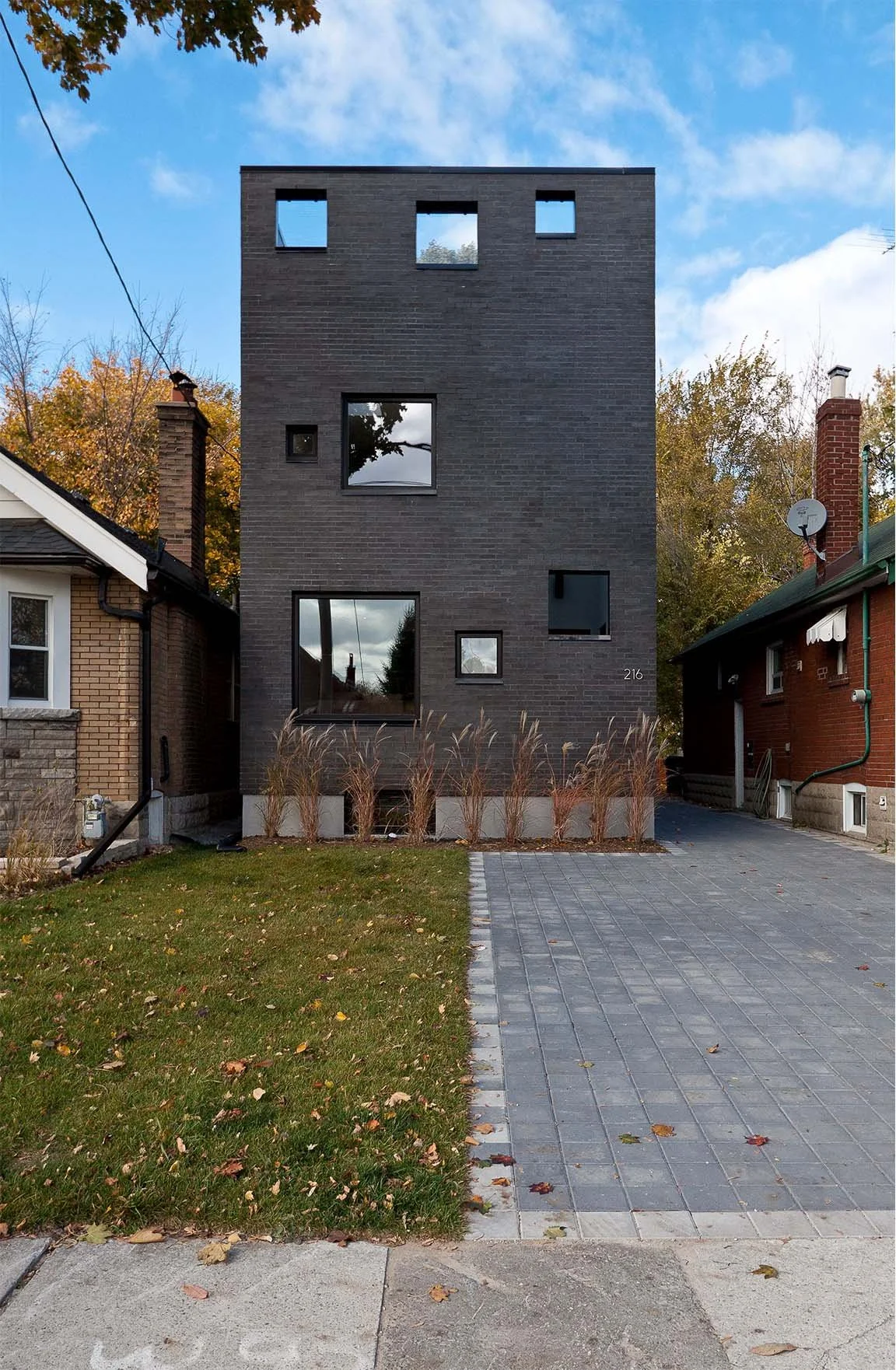 Charcoal House, a modern luxury custom home in East York, Toronto, built by Maxamin Homes, featuring a narrow three-story black brick design with geometric windows, situated between two traditional homes with a small front yard and leafless trees.