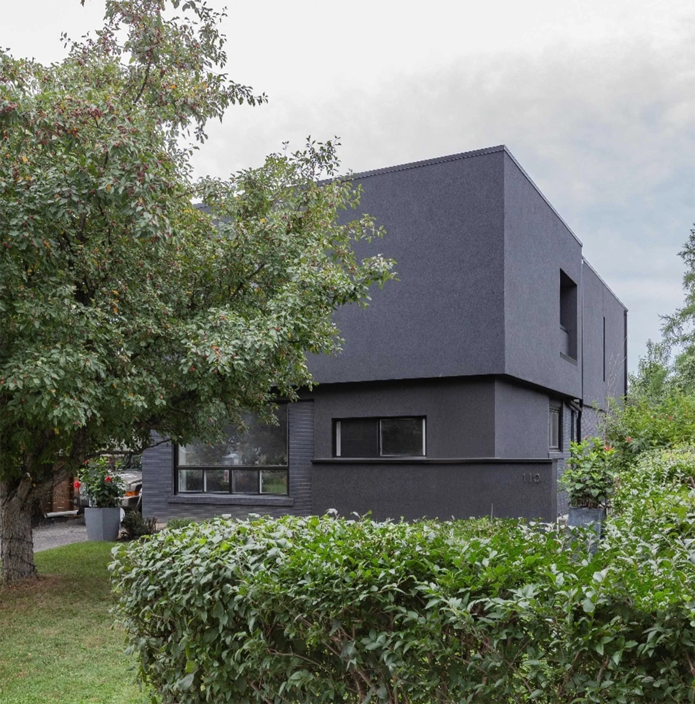 Ebenos House, a modern luxury home addition and renovation in North York, Toronto, built by Maxamin Homes, featuring a dark gray two-story boxy design with large windows, surrounded by trees and green bushes under a cloudy sky.