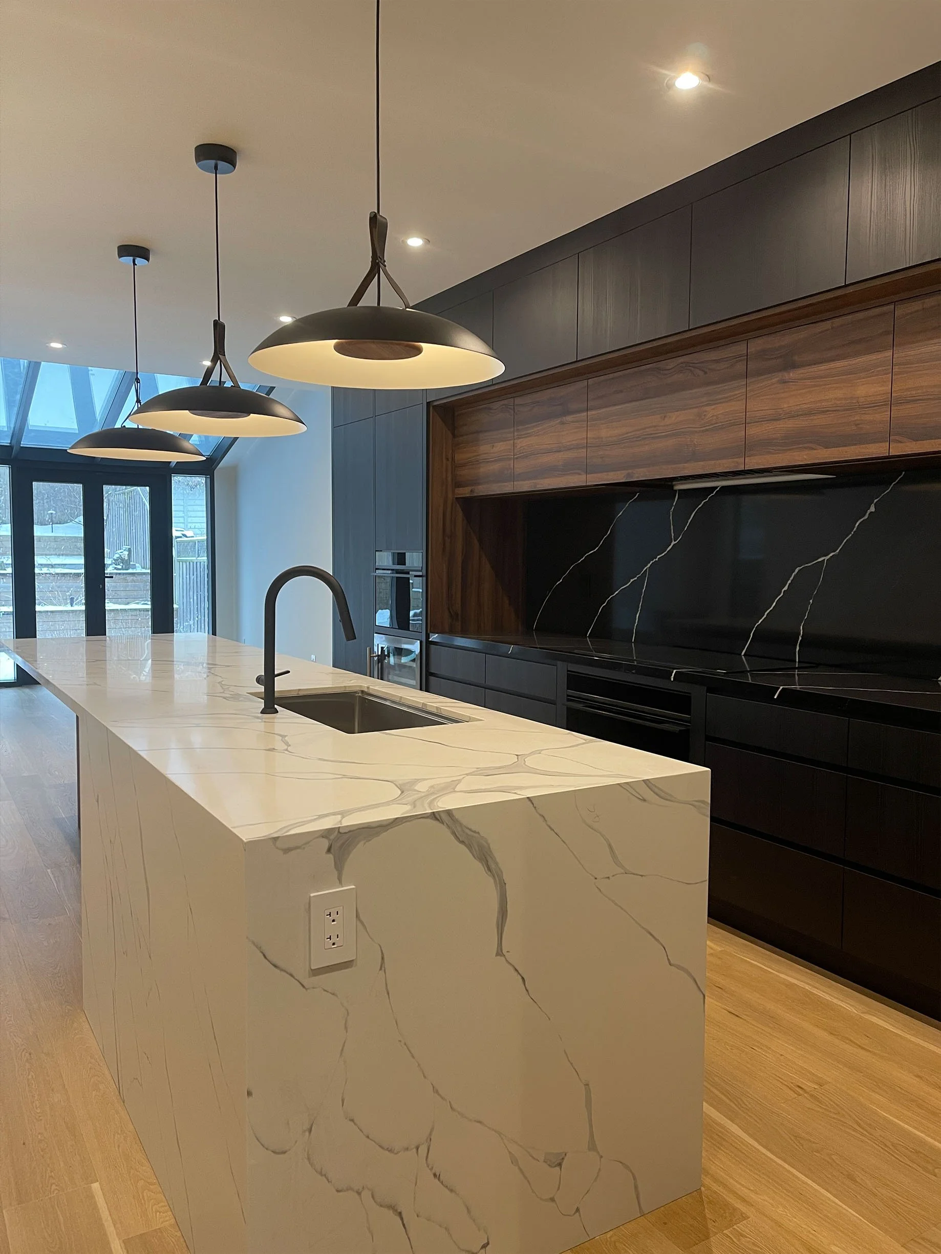 Modern kitchen with a white marble island, black sink, black and wooden cabinetry, hanging pendant lights, and large windows.