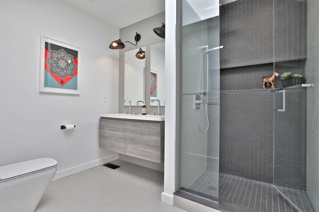 Modern bathroom with a wall-mounted vanity, two mirrors, art on the wall, a toilet, and a glass-enclosed shower with dark tiles.
