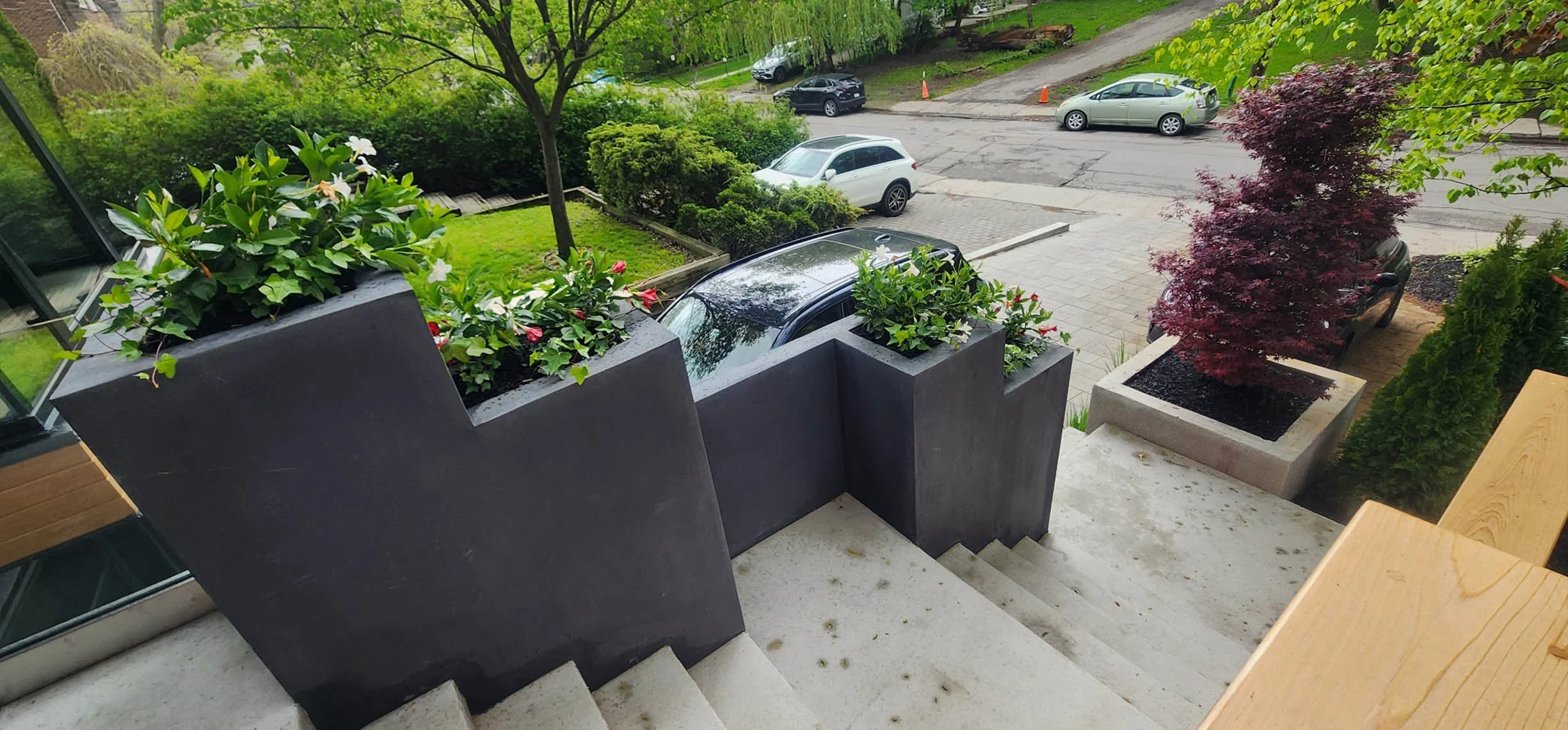 View from a staircase looking onto a sidewalk, outdoor parking, and trees. There are large rectangular planters with green plants and flowers on the staircase landing, and parked cars elsewhere. The street has several vehicles and orange traffic cone