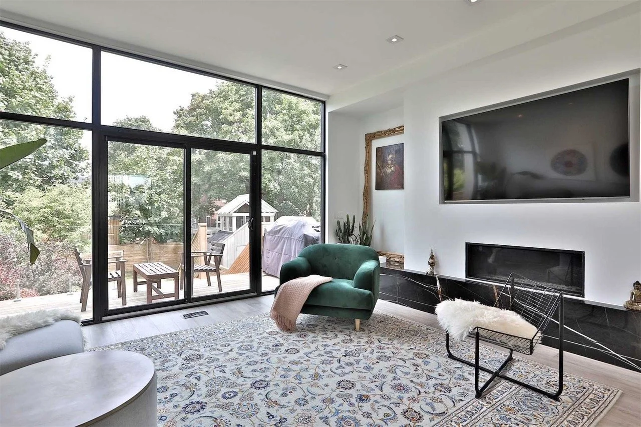 Modern living room with large glass sliding doors leading to a patio, featuring a green armchair with a beige throw blanket, a white furry bench, a patterned rug, and a wall-mounted TV