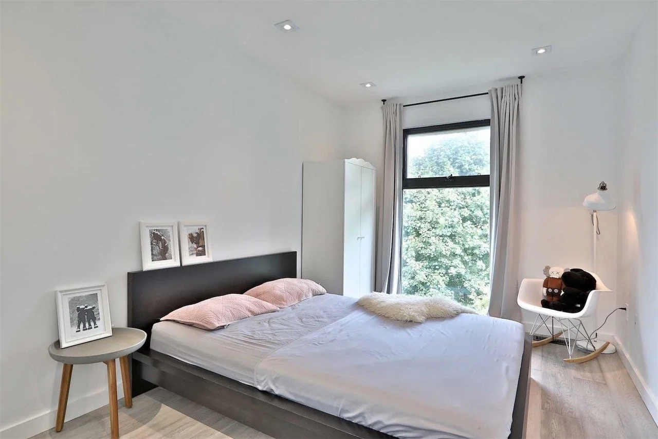 A minimalist bedroom with a bed, pink pillows, and a white fur throw. A small side table with photos, a white wardrobe, a white rocking chair with stuffed toys, and a large window with curtains overlooking trees.