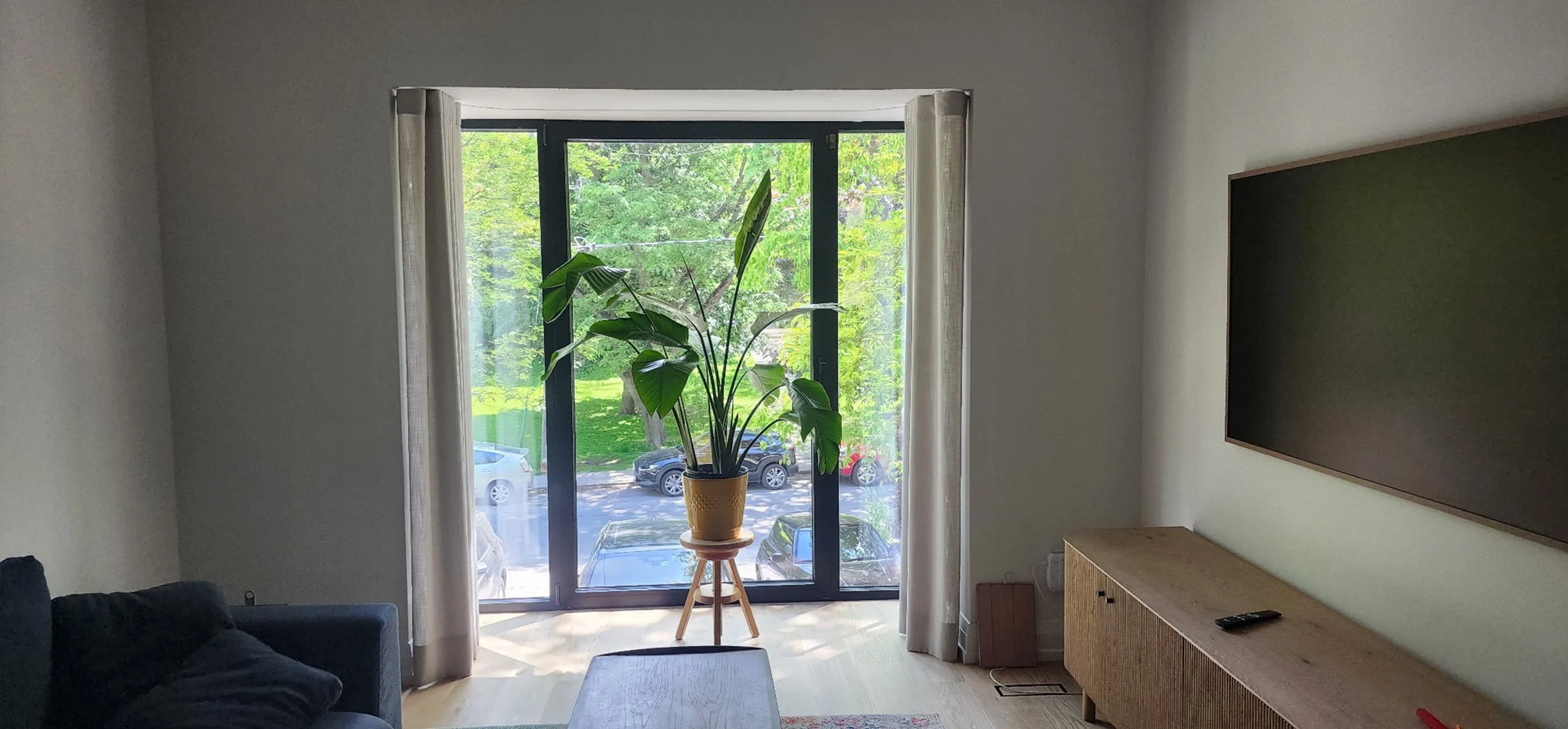 A living room with a large window and open curtains, a potted plant on a stool, a dark sofa with cushions on the left, a wooden cabinet with a remote control on top on the right, and a blank wall mounted with a large black rectangle