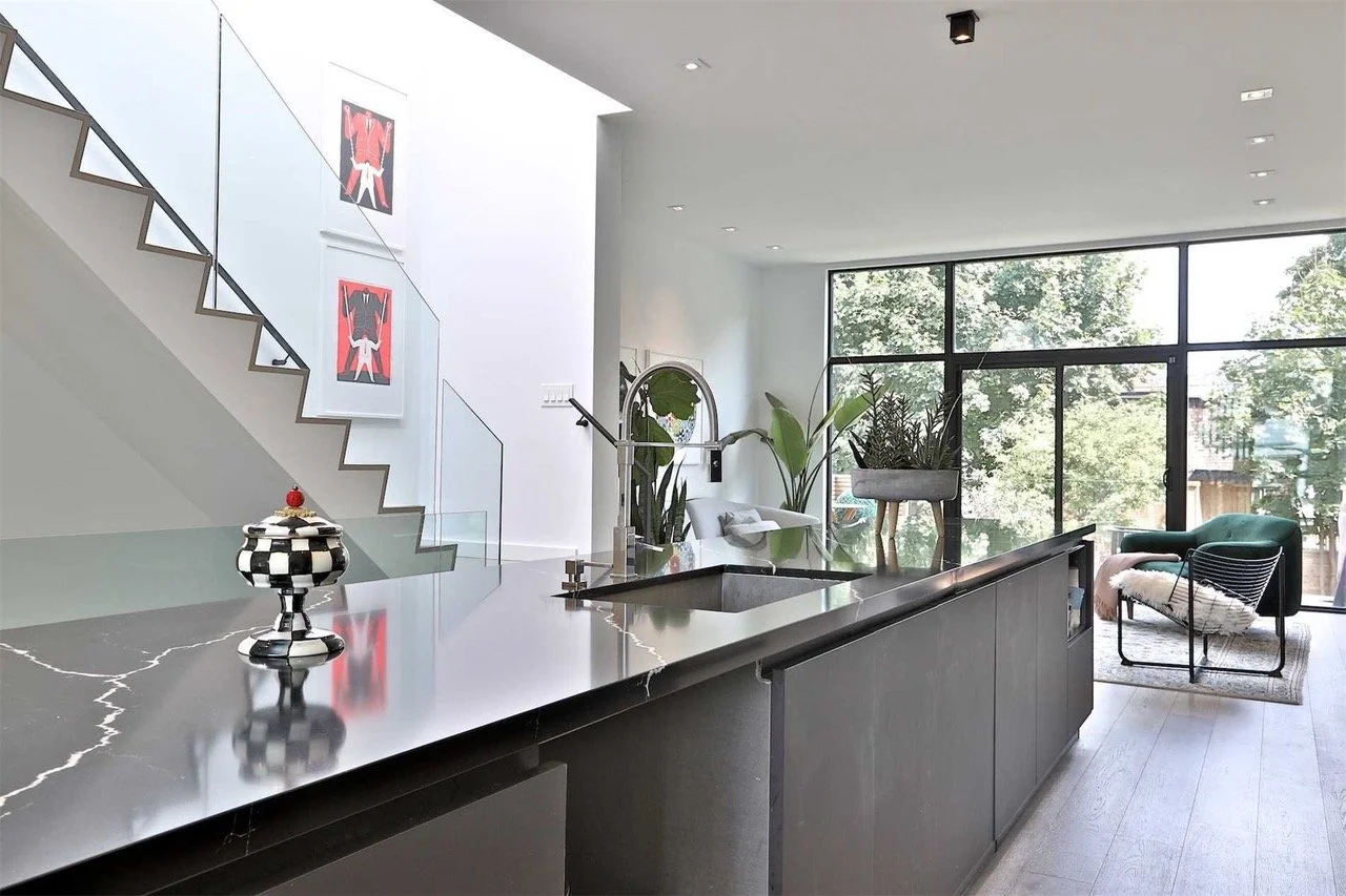 Modern open-concept living space with kitchen island, large window with trees outside, green chairs, and decorative plants.