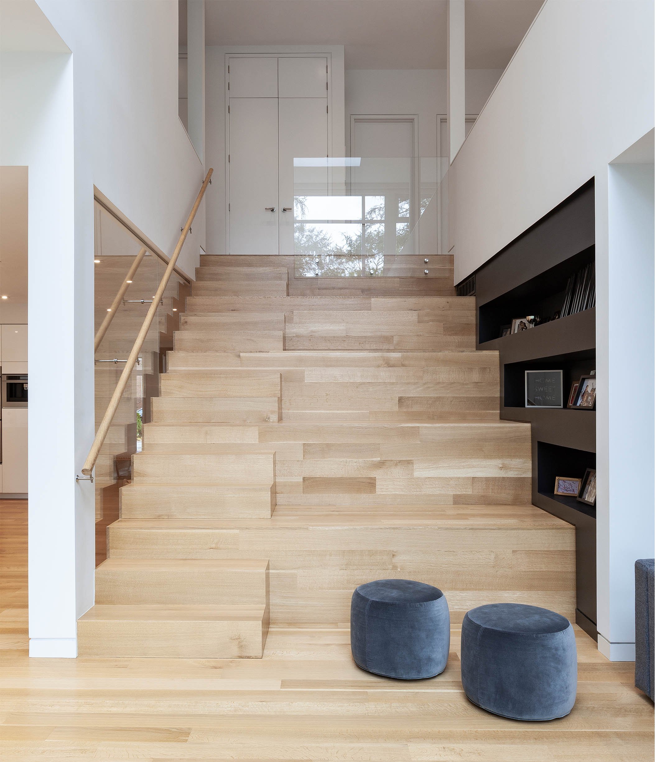 Fun House interior in East York, Toronto, built by Maxamin Homes, with wooden staircase, glass railing, black photo shelf, and blue ottomans.