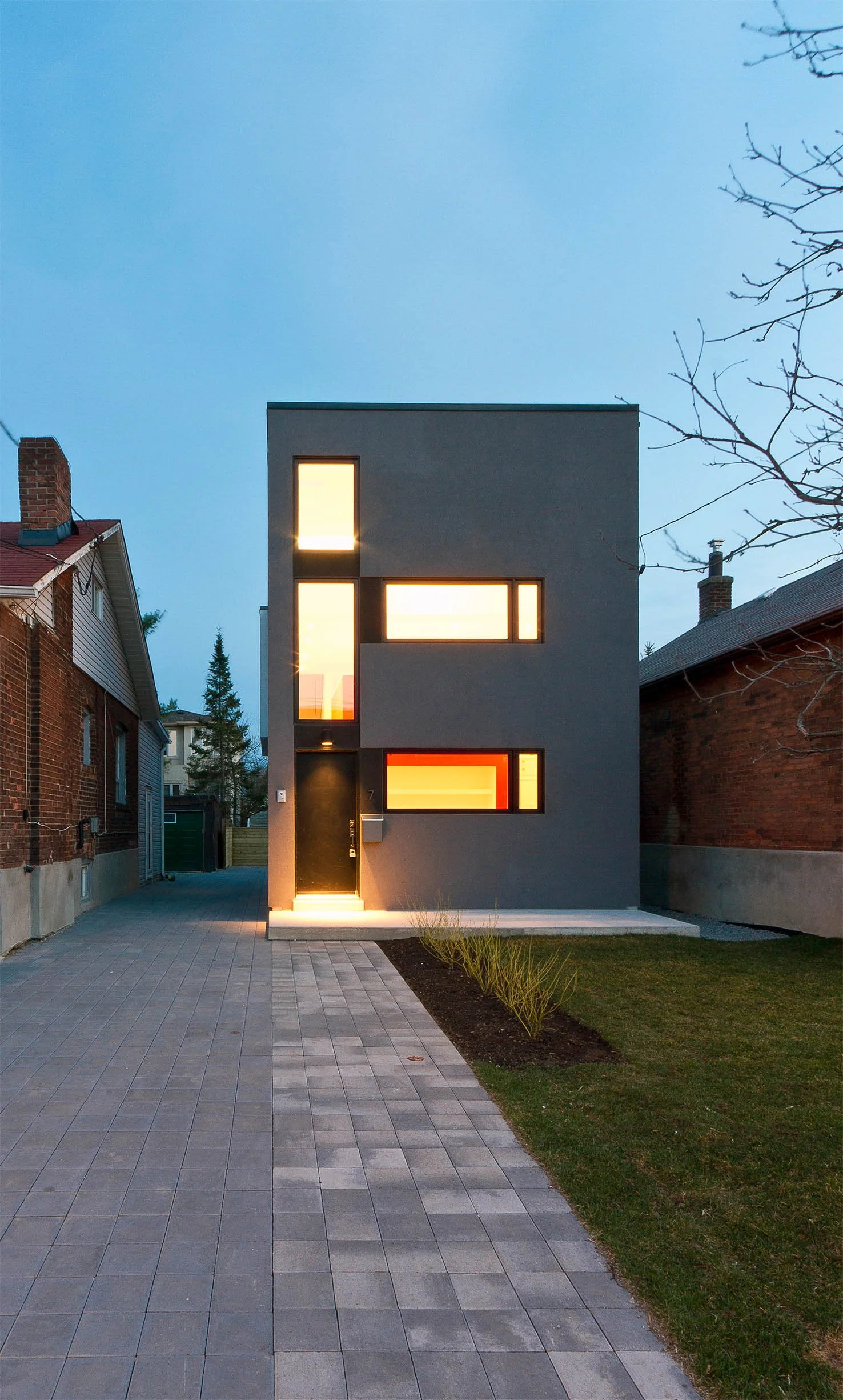 Whale House, a modern luxury custom home in East York, Toronto, built by Maxamin Homes, featuring a gray multi-story design with large rectangular windows, a black front door, and a small front garden situated between two older brick houses.