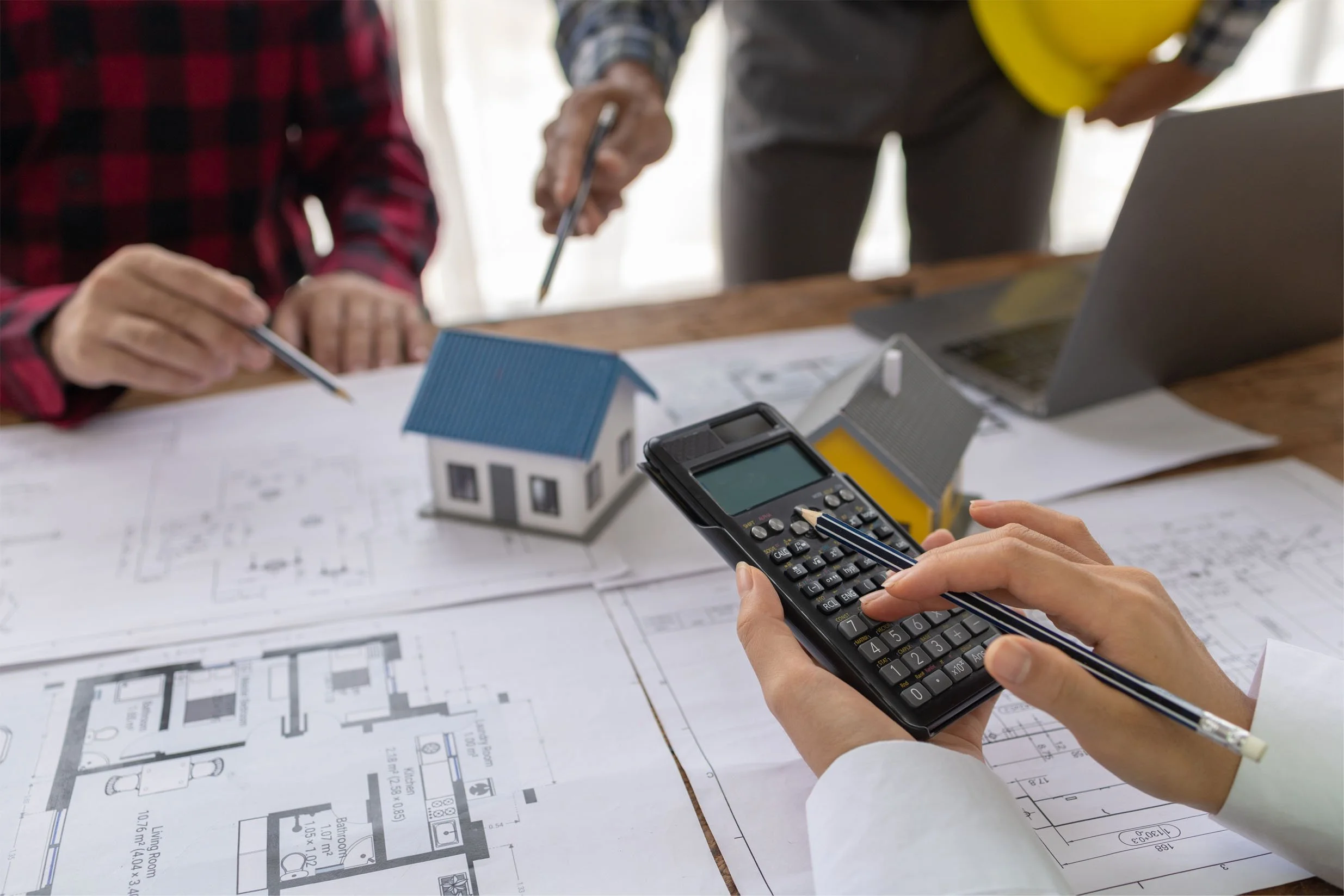 People reviewing home plans at a table with a model house, blueprints, a calculator, a laptop, and a yellow hard hat.”