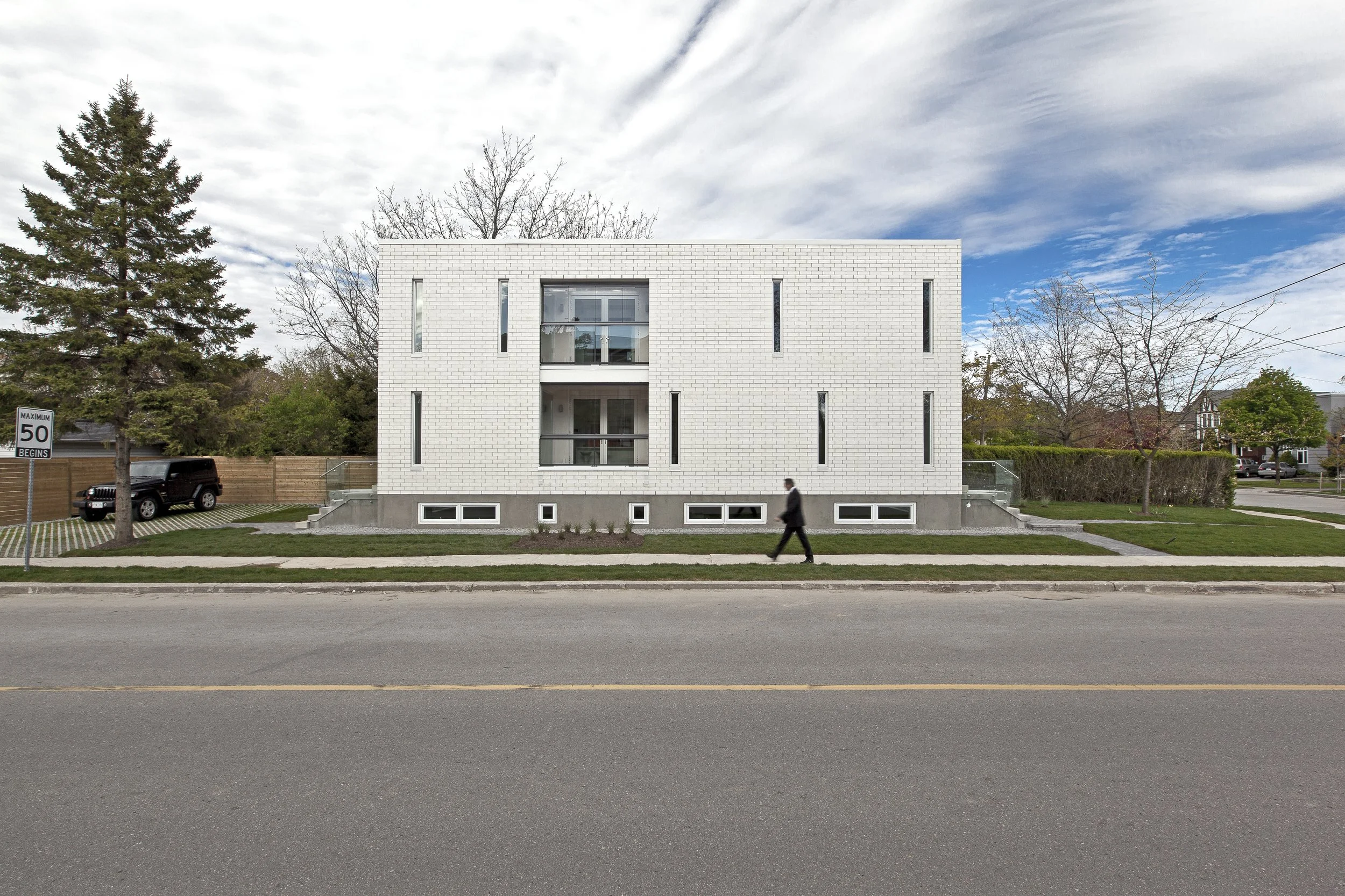 Patio House, a modern luxury custom home in North York, Toronto, built by Maxamin Homes, featuring a white brick exterior with tall narrow windows, small basement windows, and a clean streetscape with trees, parked cars, and a cloudy sky.