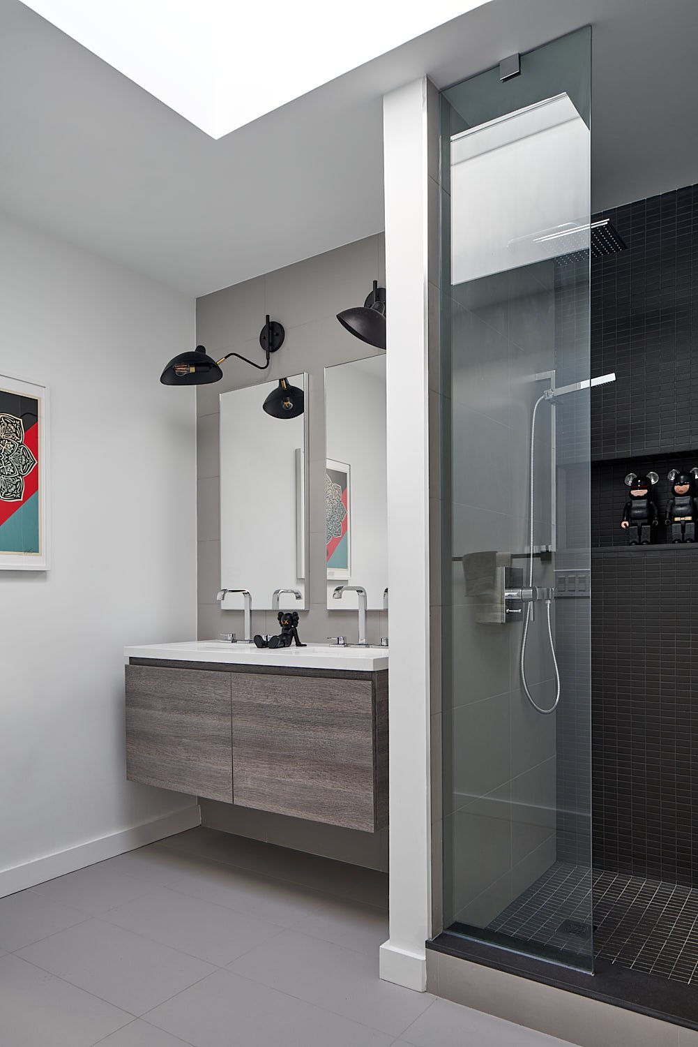 Modern bathroom with a floating wooden vanity, double sinks, large mirrors, black wall-mounted lamps, and a black-tiled shower area with a glass door.