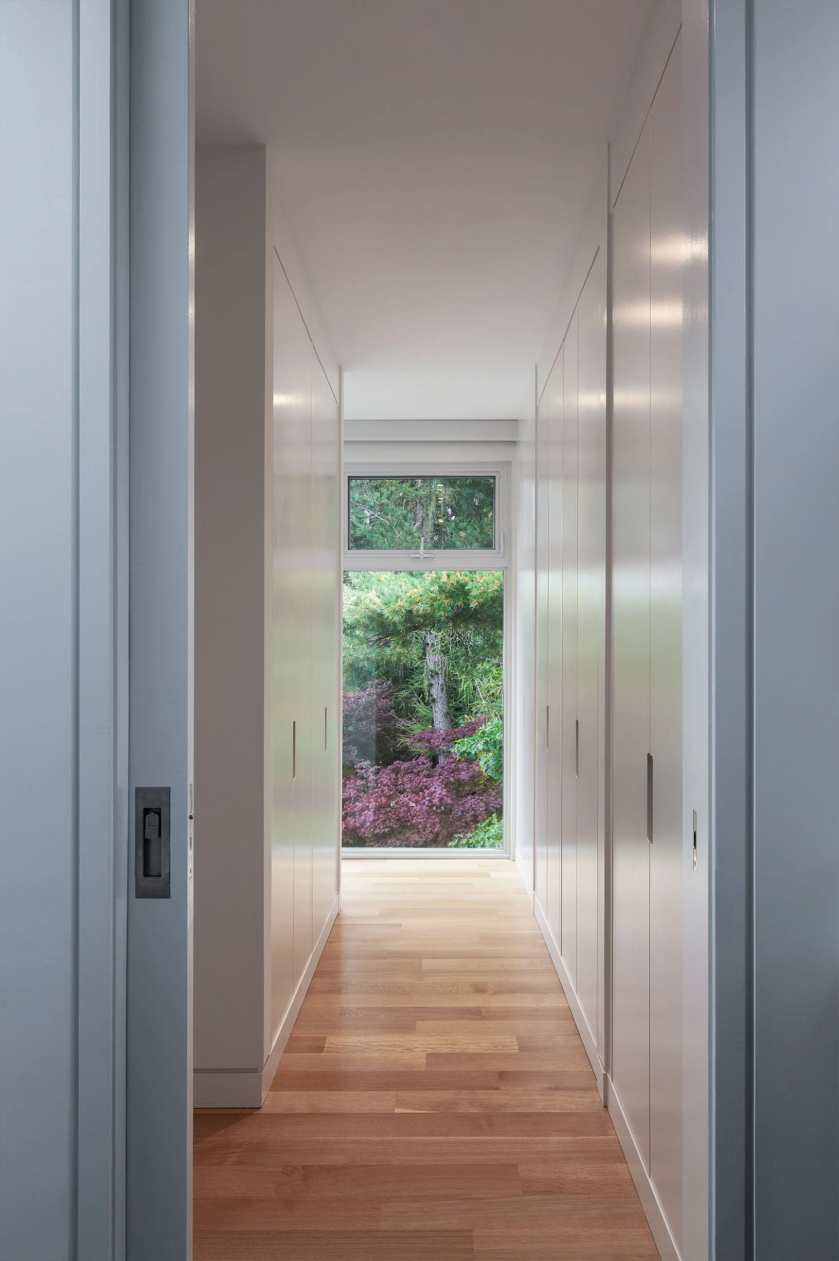 Fun House hallway in East York, Toronto, built by Maxamin Homes, with white walls, wood floors, and garden view framed by trees and pink flowers.