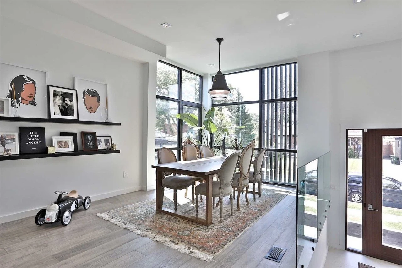 Modern dining room with a wooden table, six upholstered chairs, large windows, potted plants, a hanging pendant light, patterned rug, framed artwork, and a toy car on light-colored flooring.