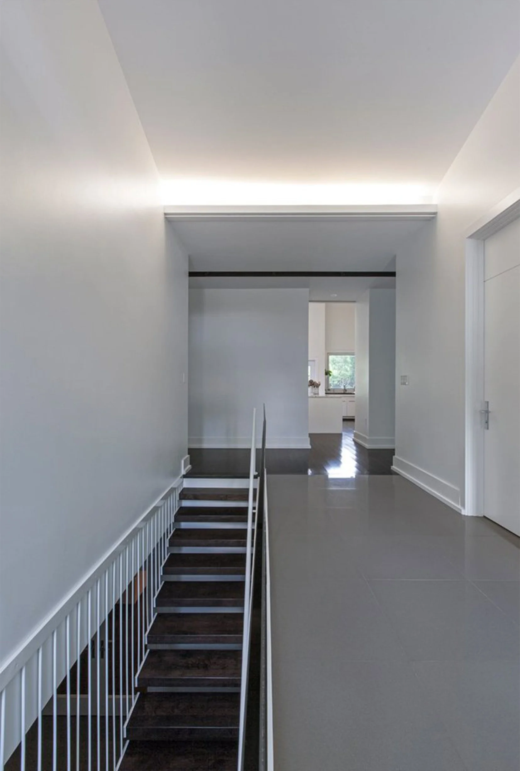 Indoor hallway with staircase leading downstairs, white walls, and modern lighting. View into a bright room with large window.
