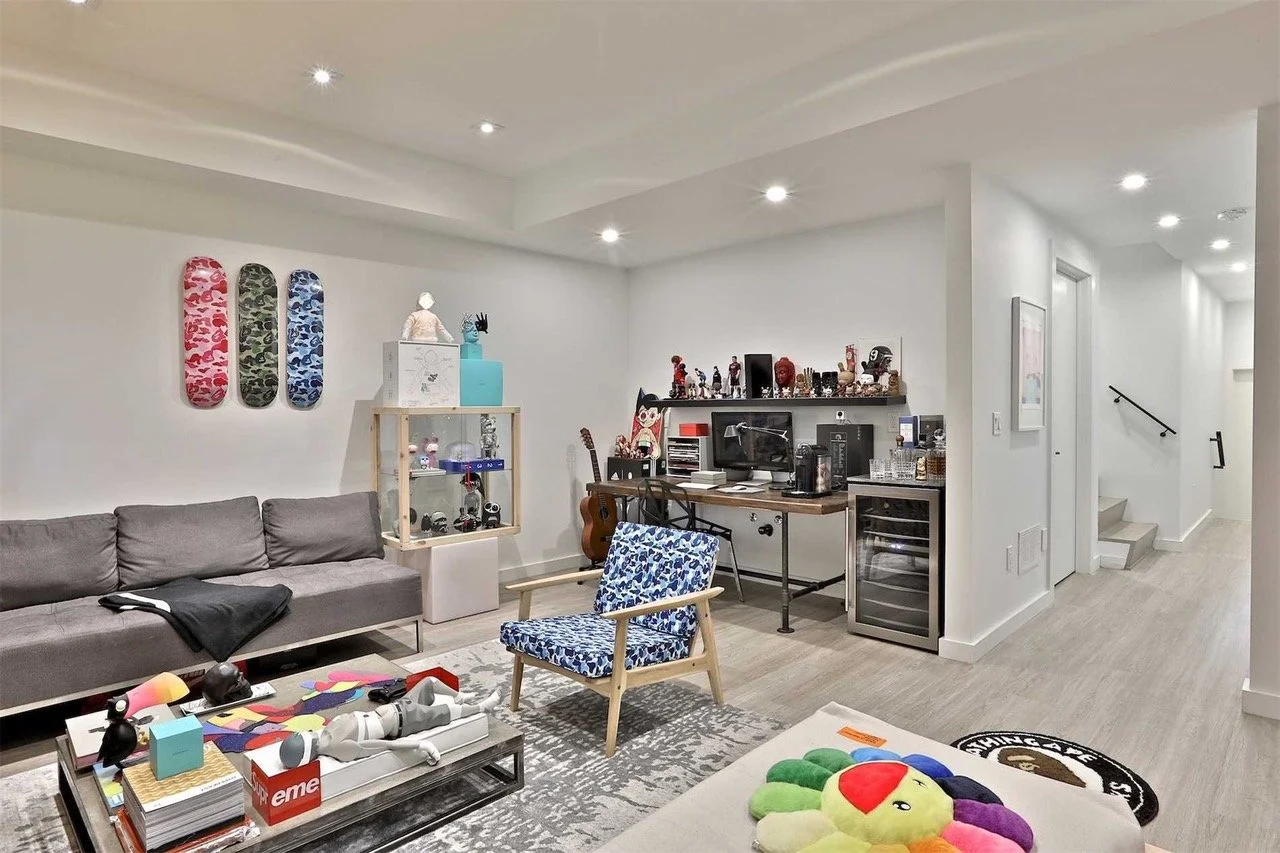 Living room with gray sofa, blue patterned armchair, coffee table with toys, skateboard decorations on the wall, and a desk with computer and collectibles. Stairs leading upstairs are visible in the background.