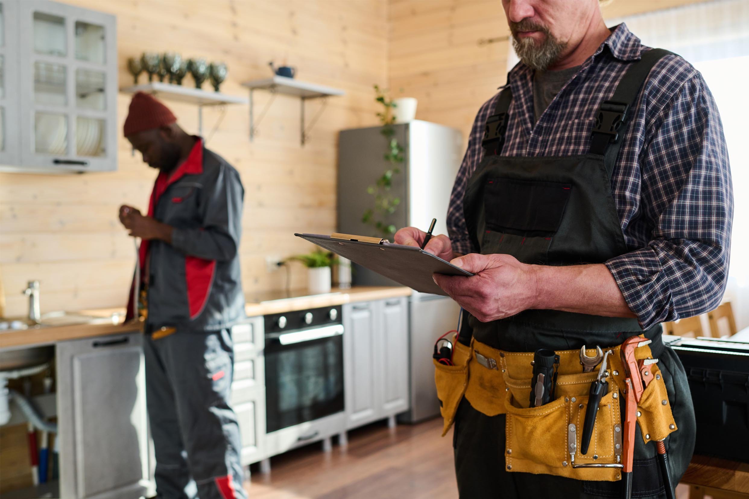 A man wearing a tool belt and plaid shirt holding a clipboard, standing in a kitchen, with a woman in work clothes in the background.