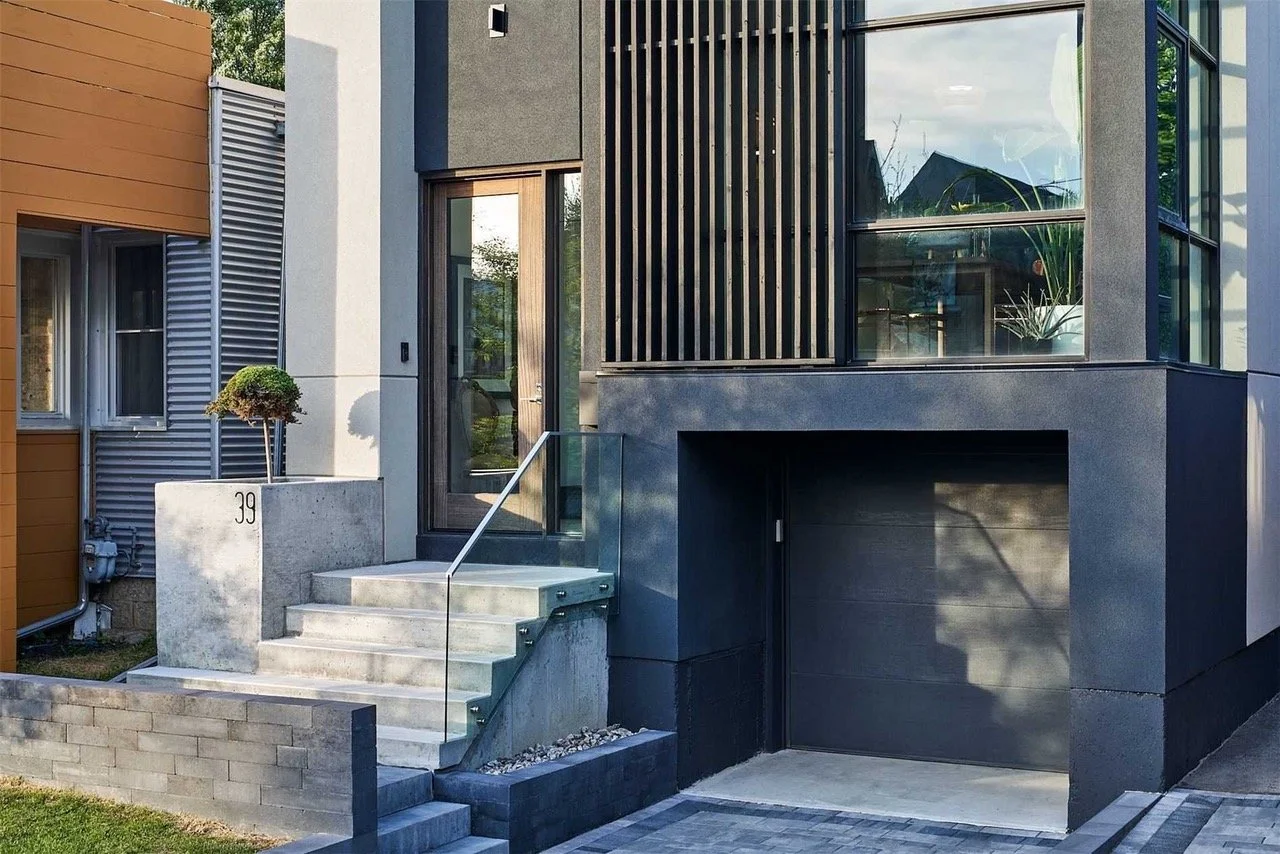 Modern house facade with concrete stairs, large glass windows, and a garage door on the lower level.