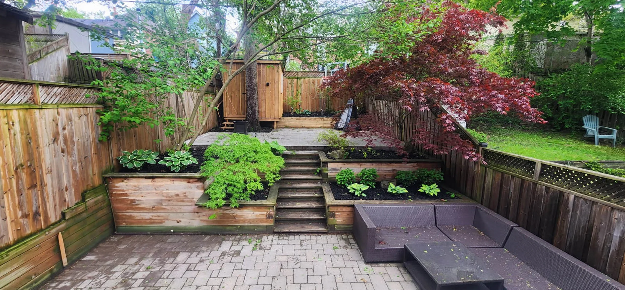 Backyard garden with wooden fences, raised garden beds with plants, a small wooden shed, a stone patio, a gray outdoor seating area, colorful trees including a red maple, and a lawn with a blue Adirondack chair.