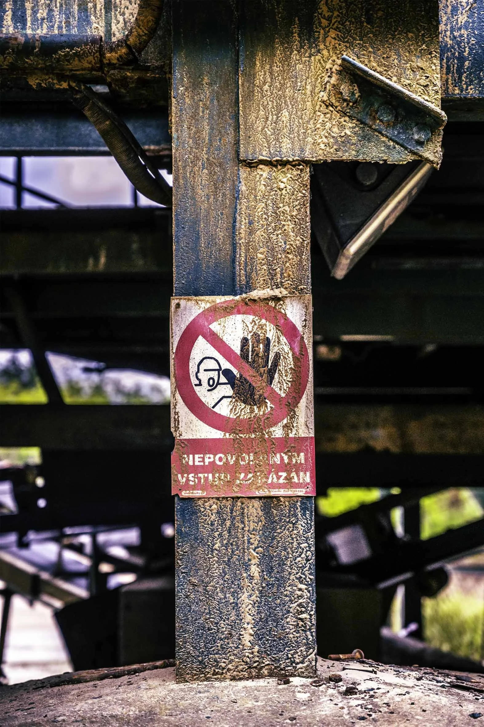 A rusted metal post with a worn, faded sign indicating no entry for safety helmets, in an industrial setting or construction site.