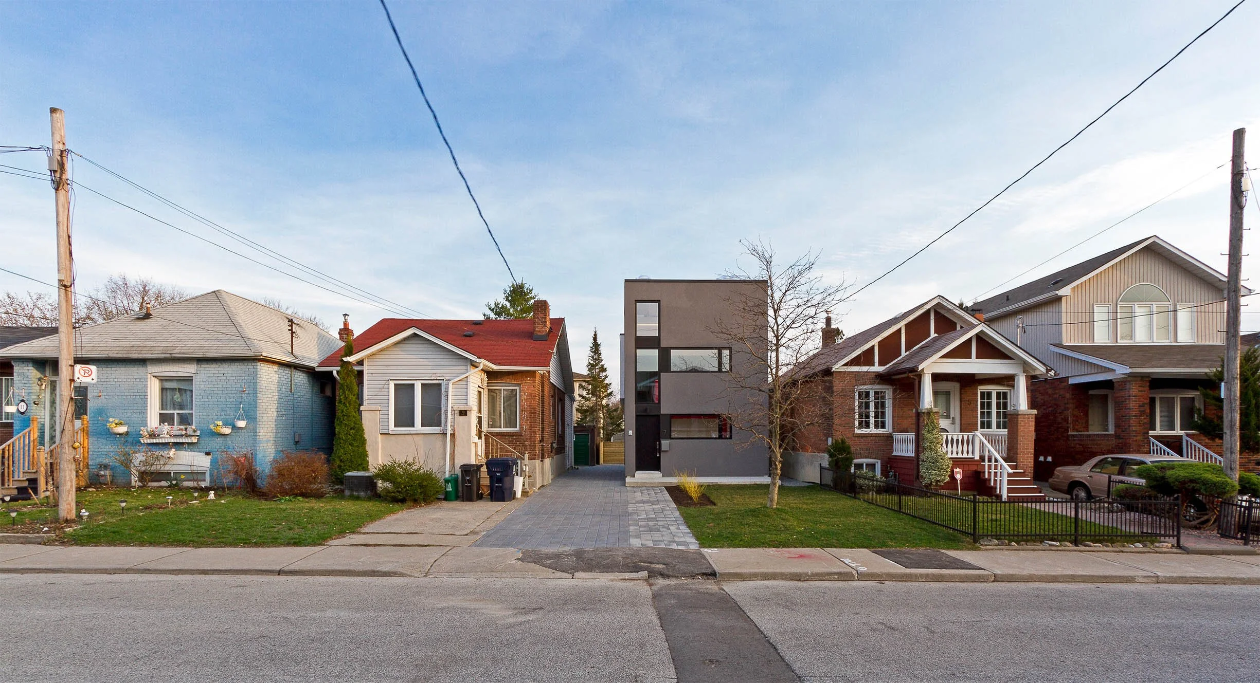 Street view near Whale House in East York, Toronto, showing modern gray home between two traditional brick houses with lawns and trees.