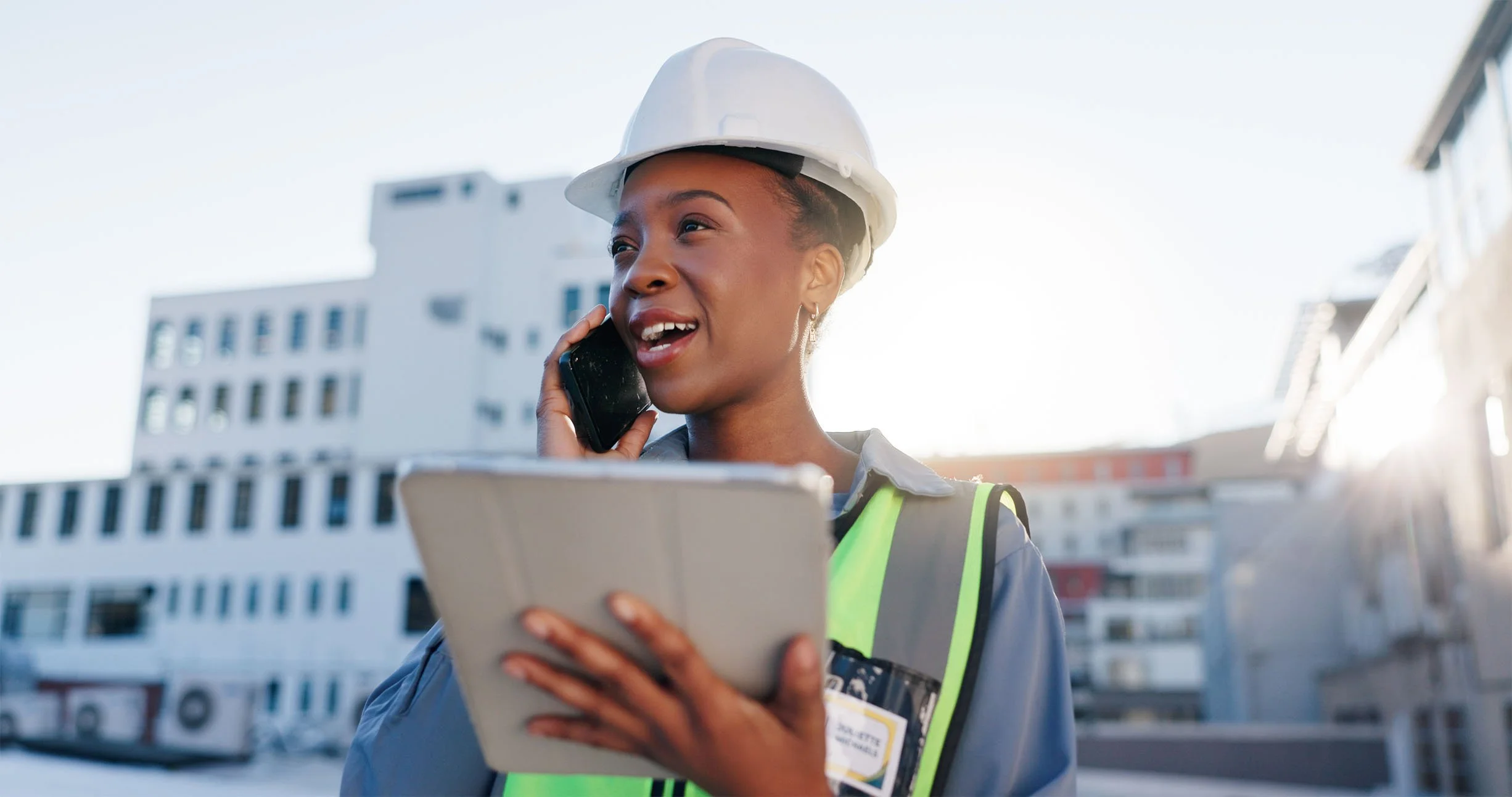 A woman in a hard hat and safety vest is talking on a phone while holding a tablet, standing outdoors with buildings in the background.