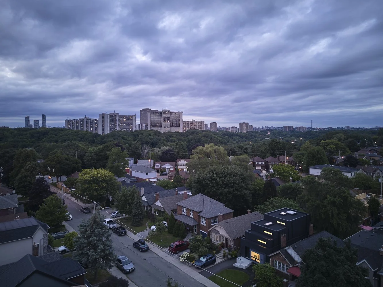 Urban neighborhood view near Periscope House in East York, Toronto, showing family homes, trees, and distant high-rises under a cloudy sky.
