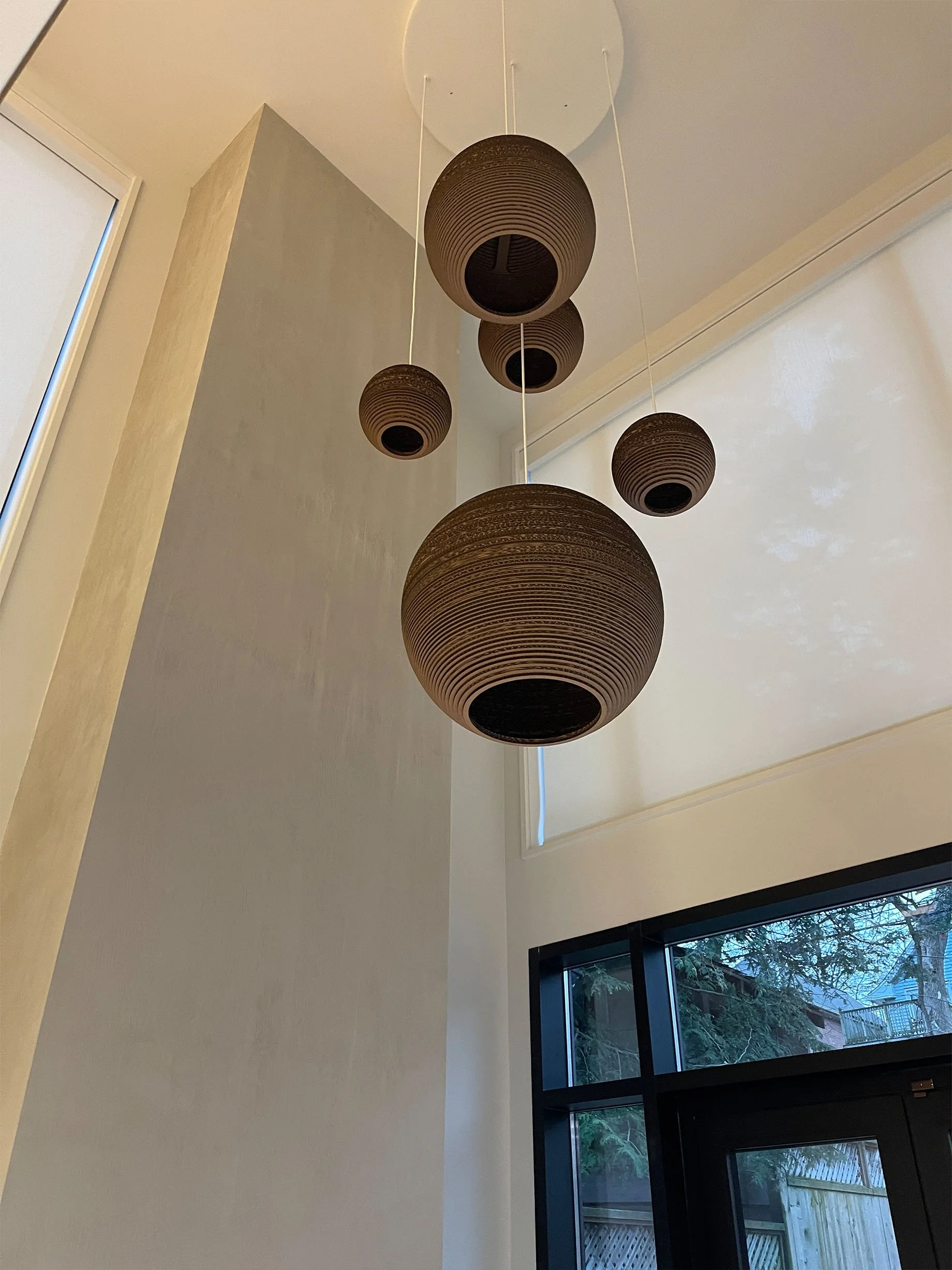 A modern interior ceiling light fixture with five dark-colored, round, ribbed pendant lamps hanging from the ceiling above a large window with a view of trees outside.