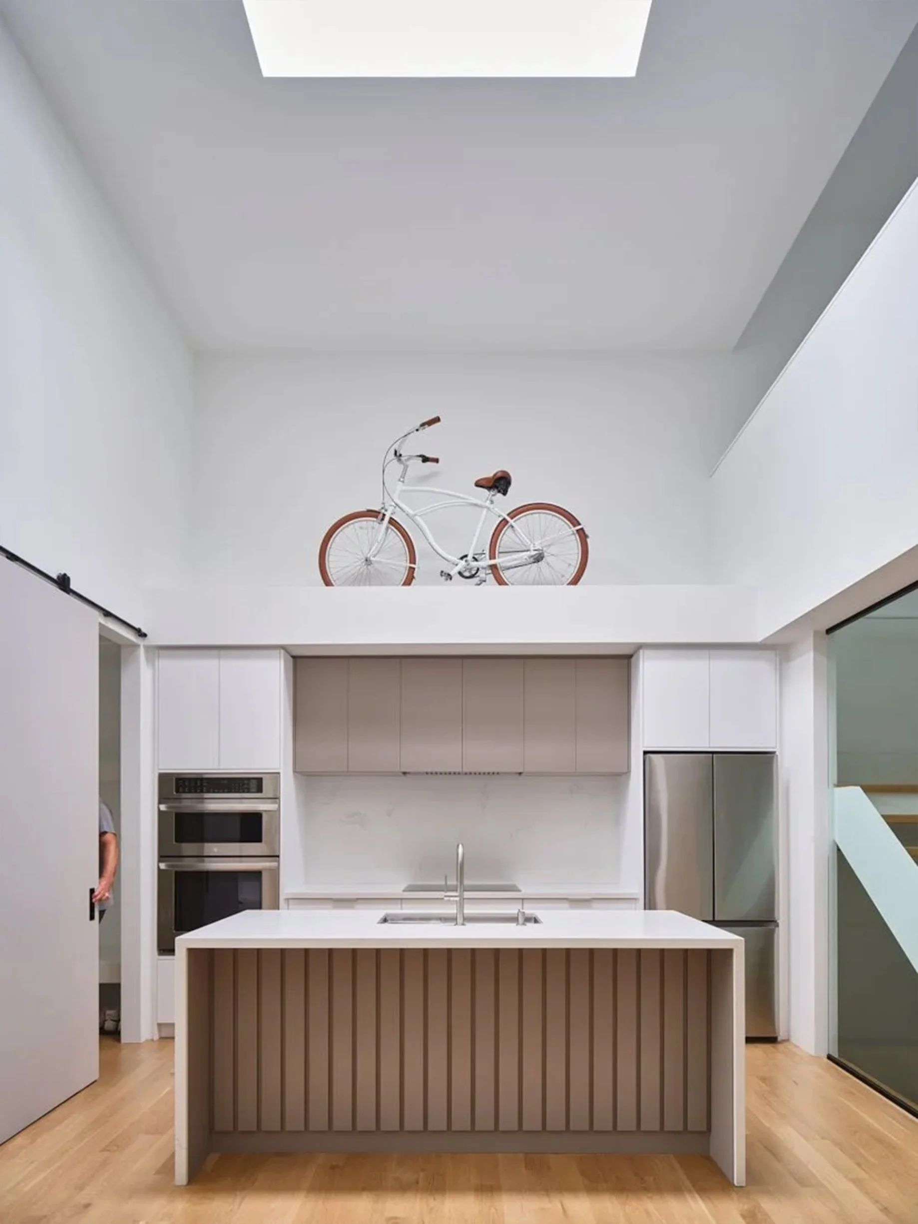 Periscope House kitchen in East York, Toronto, built by Maxamin Homes, with white island, beige cabinets, stainless ovens, and a bicycle displayed above.