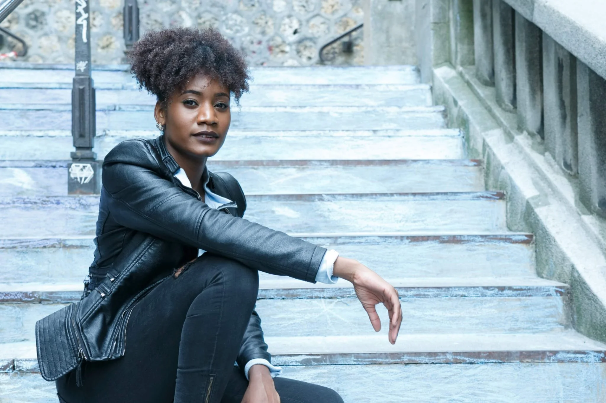 Jeune femme assise sur des escaliers en pierre, portant une veste en cuir noire, cheveux bouclés et regardant vers l'avant.