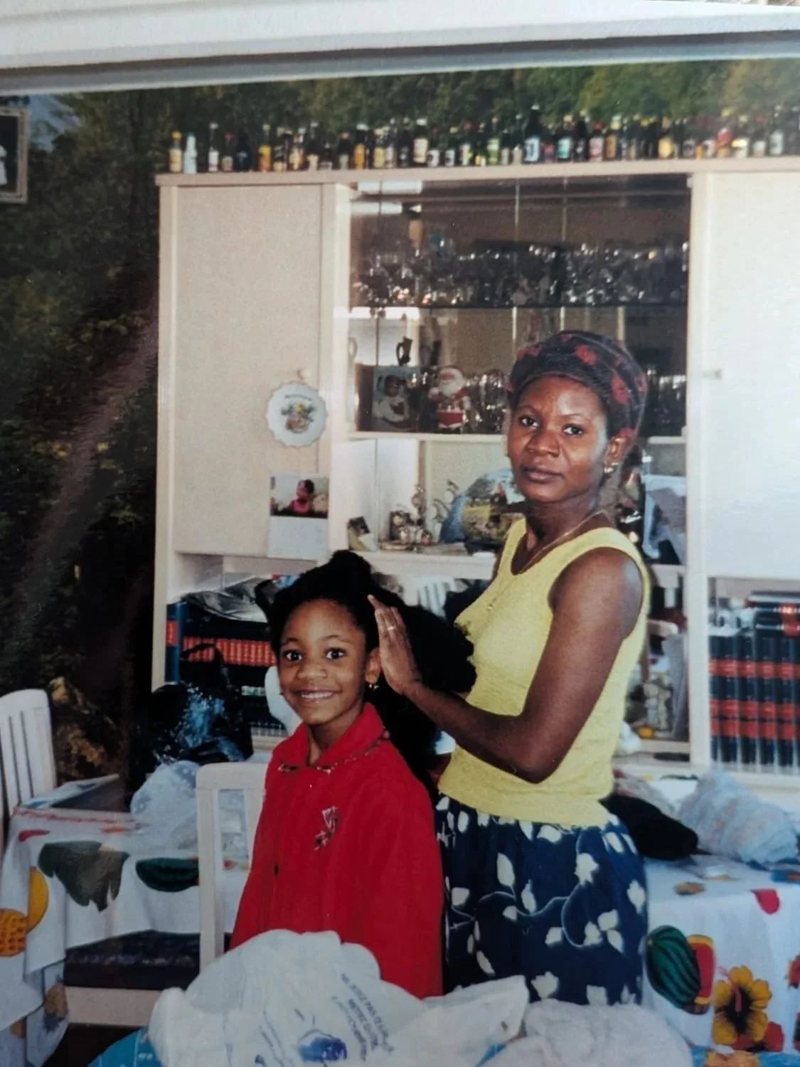 Une femme et une jeune fille posent pour une photo à l'intérieur d'une maison. La femme porte un bandeau sur la tête et un débardeur jaune, tandis que la fille porte une veste rouge. En arrière-plan, il y a une étagère avec des décorations et des livres, ainsi qu'une table avec une nappe à motifs colorés.
