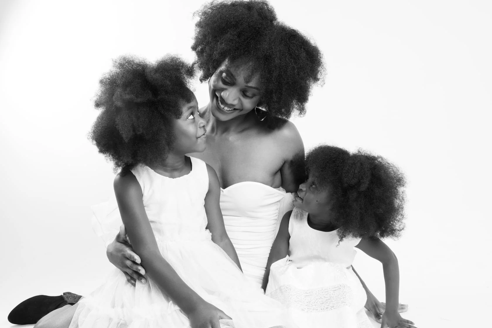Une femme avec deux jeunes filles, tous en robes blanches, souriant et partageant un moment de complicité. La femme tient les filles dans une pose affectueuse. L'image est en noir et blanc.