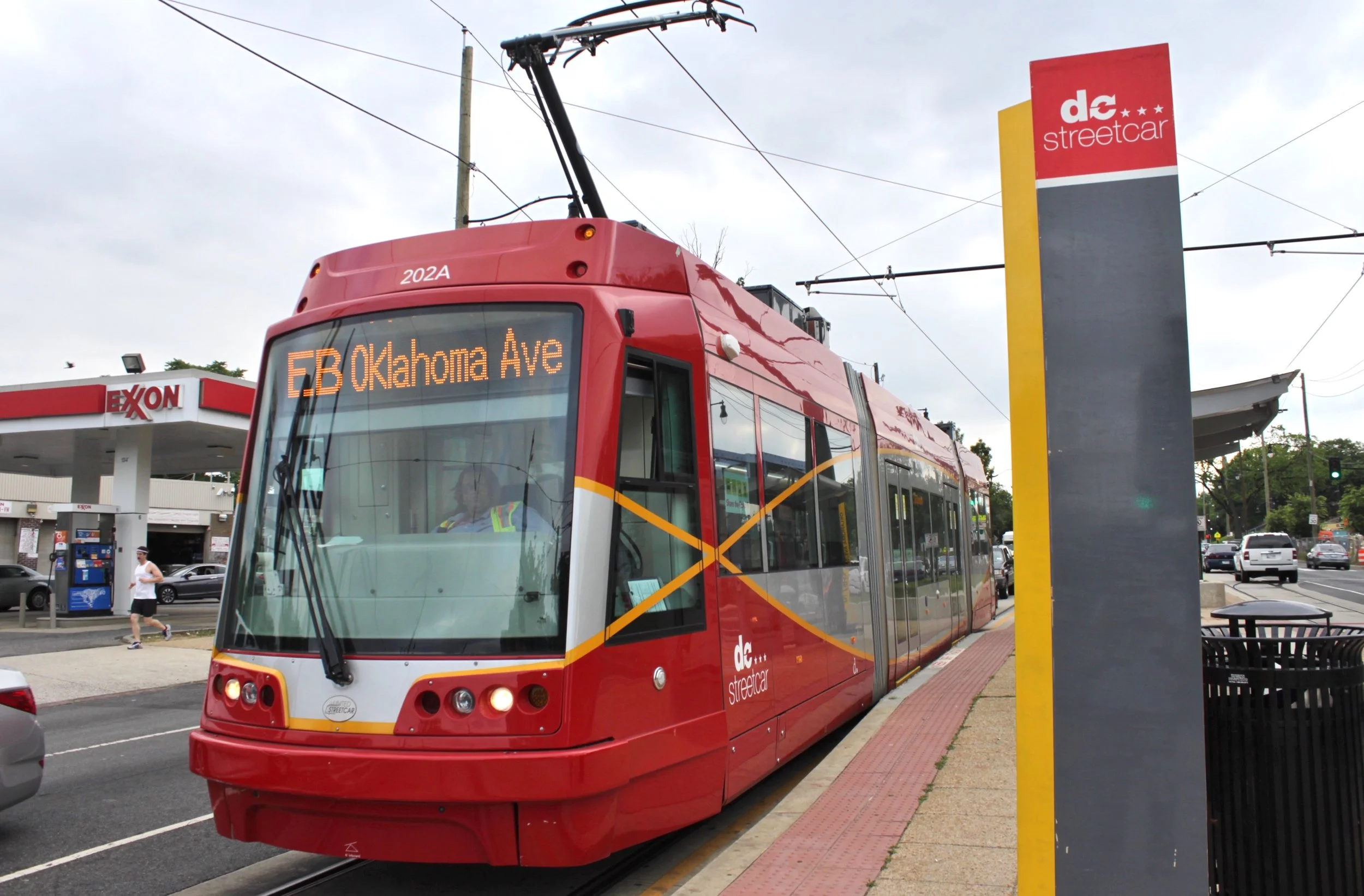 A red DC Streetcar stopped at a station marked “EB Oklahoma Ave” in Washington, D.C., with an Exxon gas station visible nearby, representing urban public transportation and neighborhood transit access