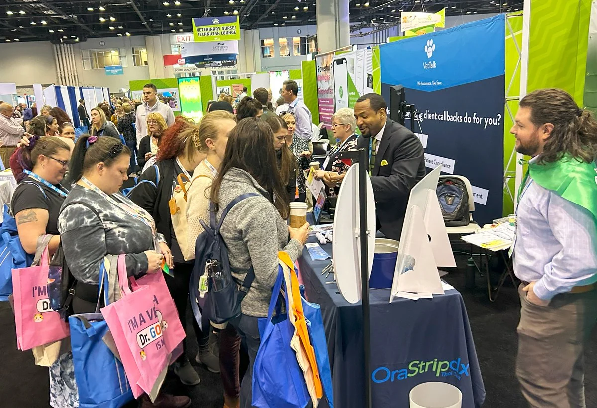 People gather around a trade show booth at a convention center, with signage for VetTalk and OraStrip in the background.