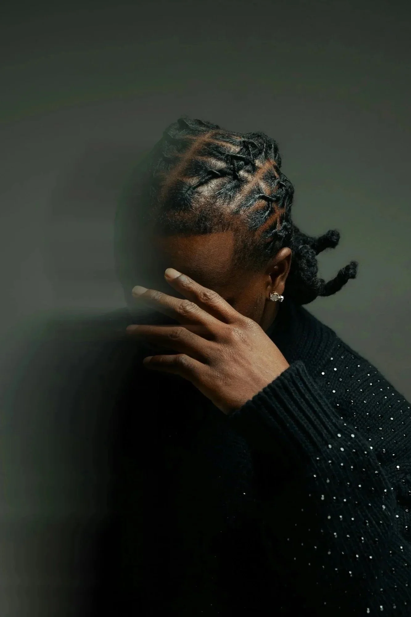 A man of color with braided hair and earrings poses with his hand near his face, wearing a black, sparkly sweater against a dark background.