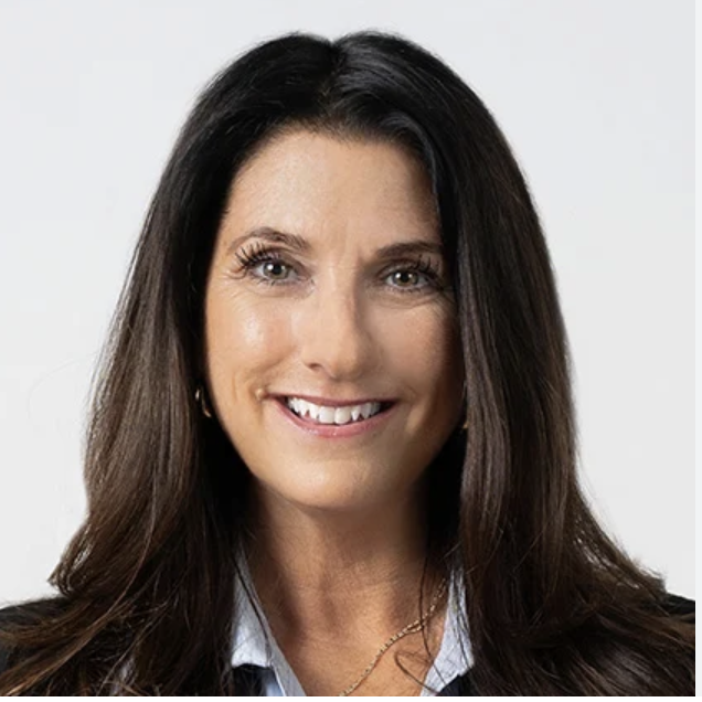A woman with long dark hair, smiling, wearing a light blue collared shirt and a dark blazer.