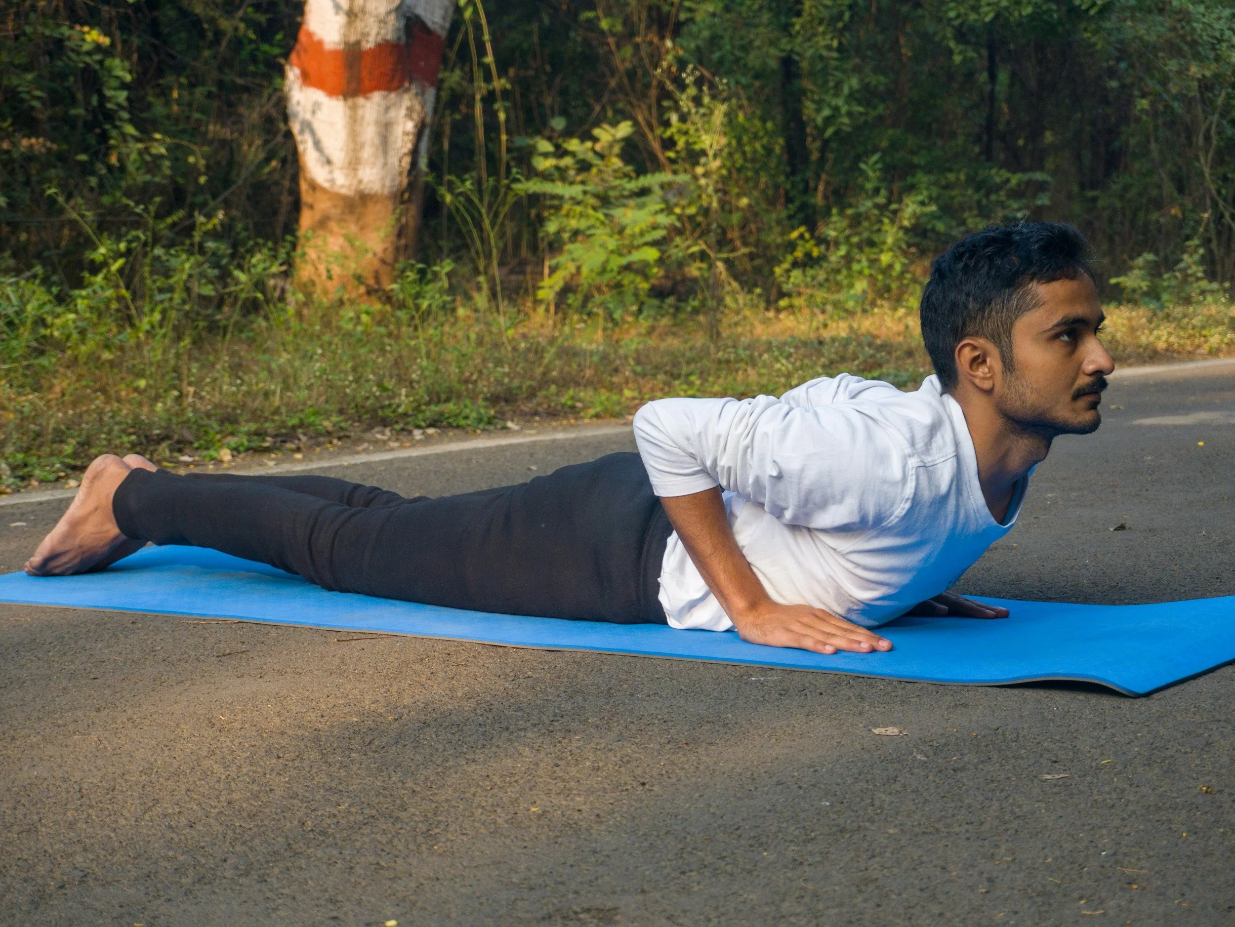 A man of color lying on a yoga mat outdoors performing a yoga pose on a paved surface next to a road, with green trees and bushes in the background.