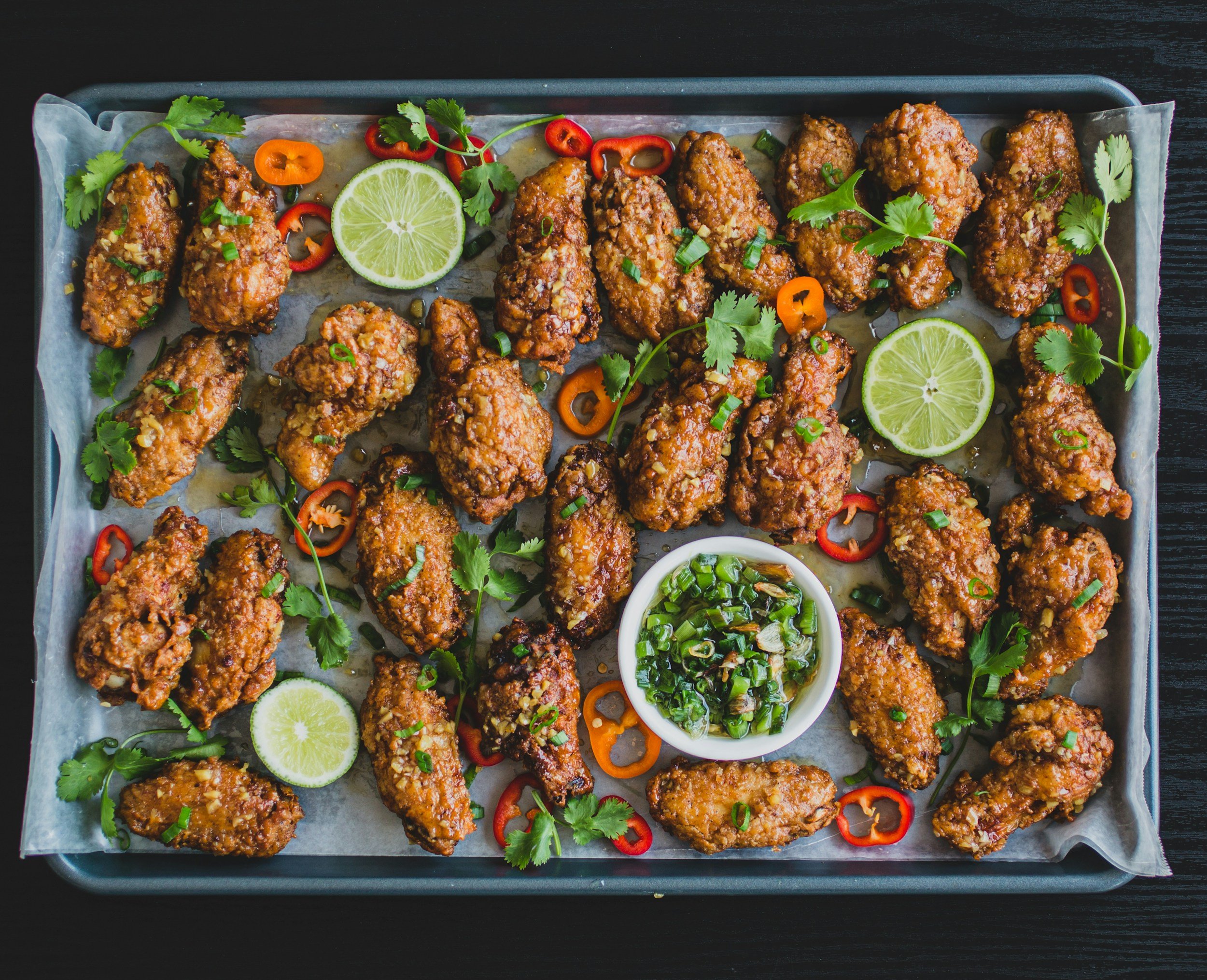 Fried chicken wings garnished with chopped green onions, cilantro, and slices of lime, with a small bowl of dipping sauce on a tray lined with parchment paper.