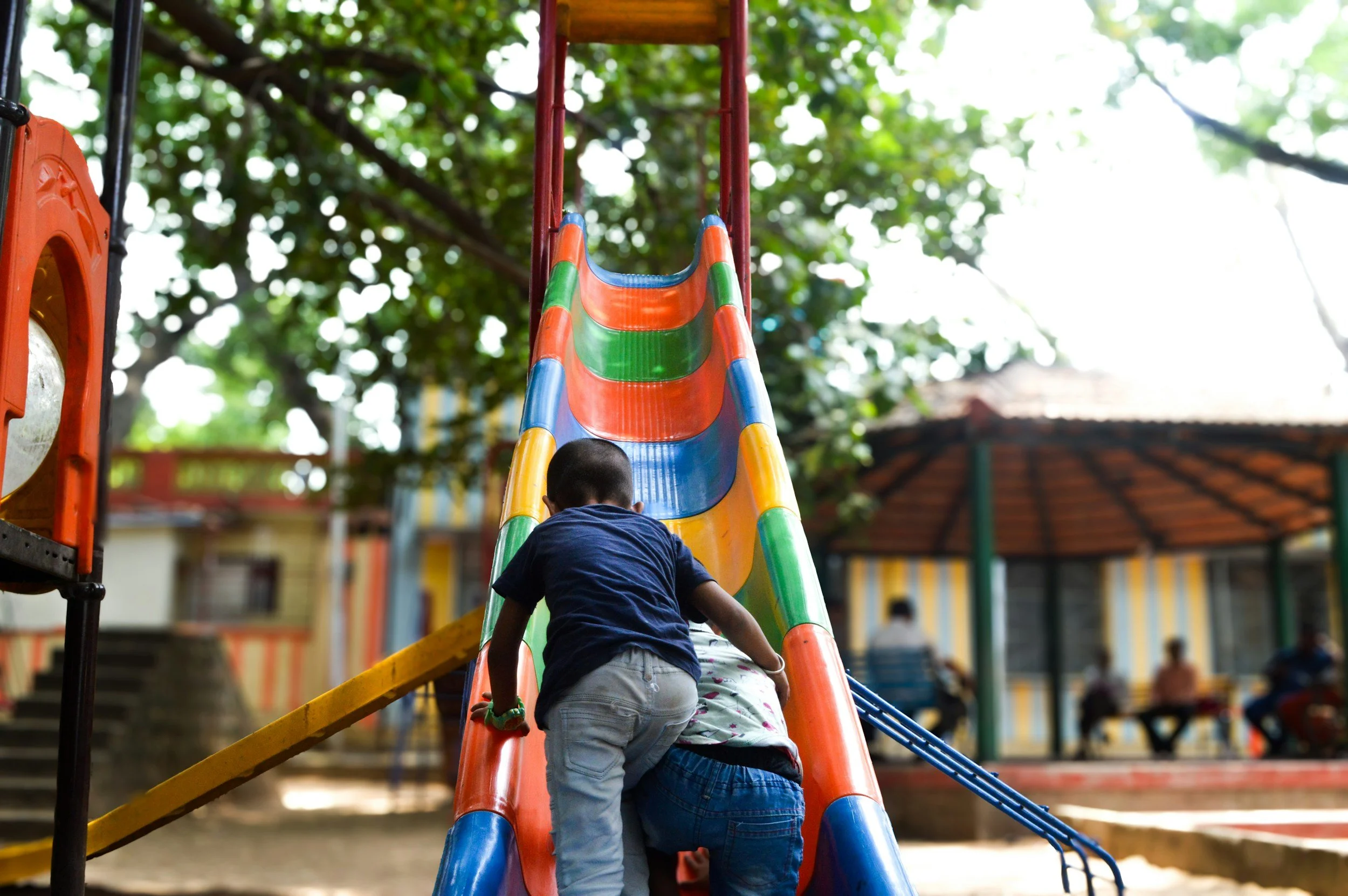 Diverse group of children climbing a colorful slide at a playground with trees and buildings in the background.