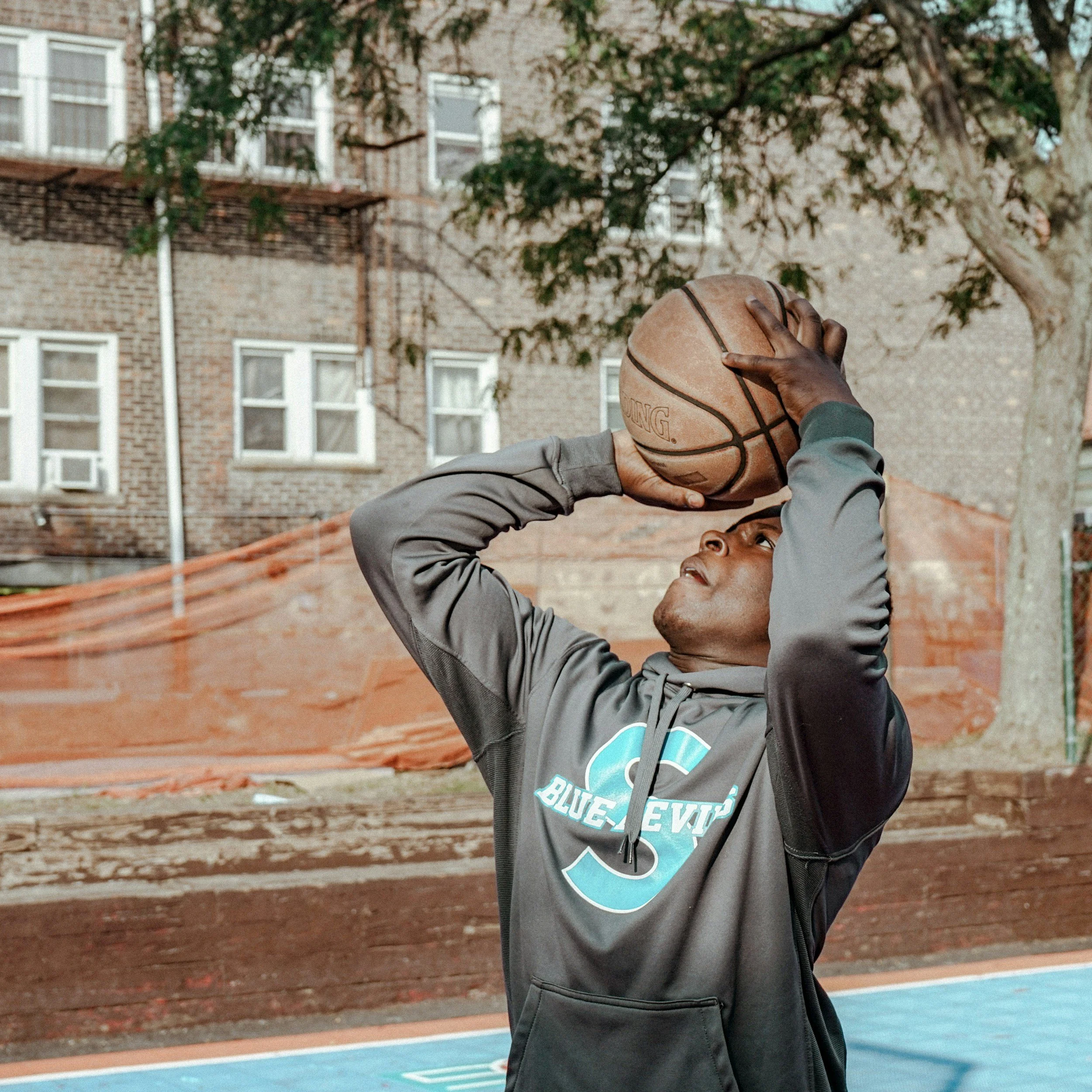A young boy of color in a gray hoodie is outdoors playing basketball, holding a basketball with one hand and preparing to shoot with the other on a court surrounded by a brick building and trees.