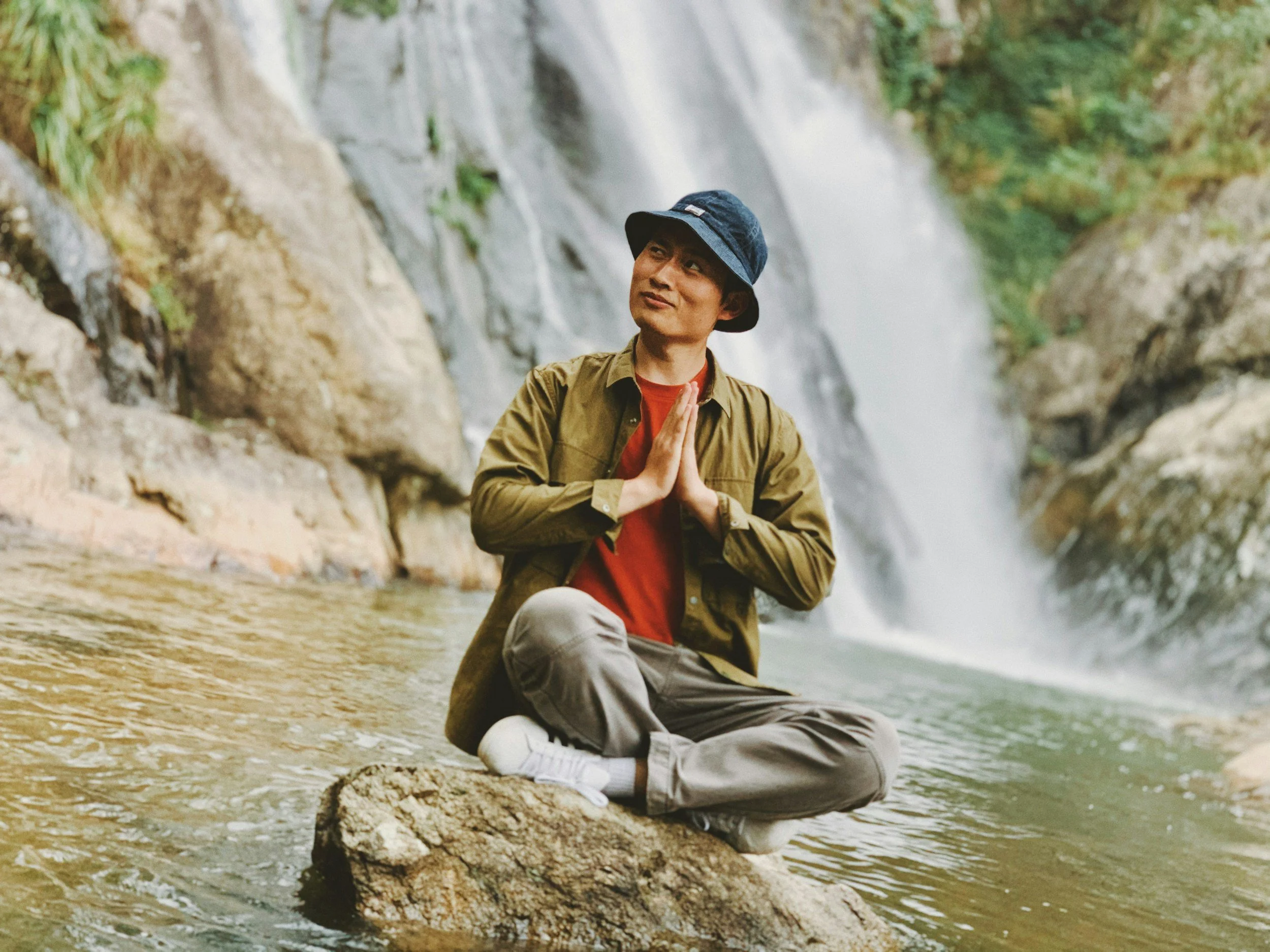 An older man of color is sitting cross-legged on a rock near a waterfall, with his hands pressed together in a prayer gesture and looking up thoughtfully.