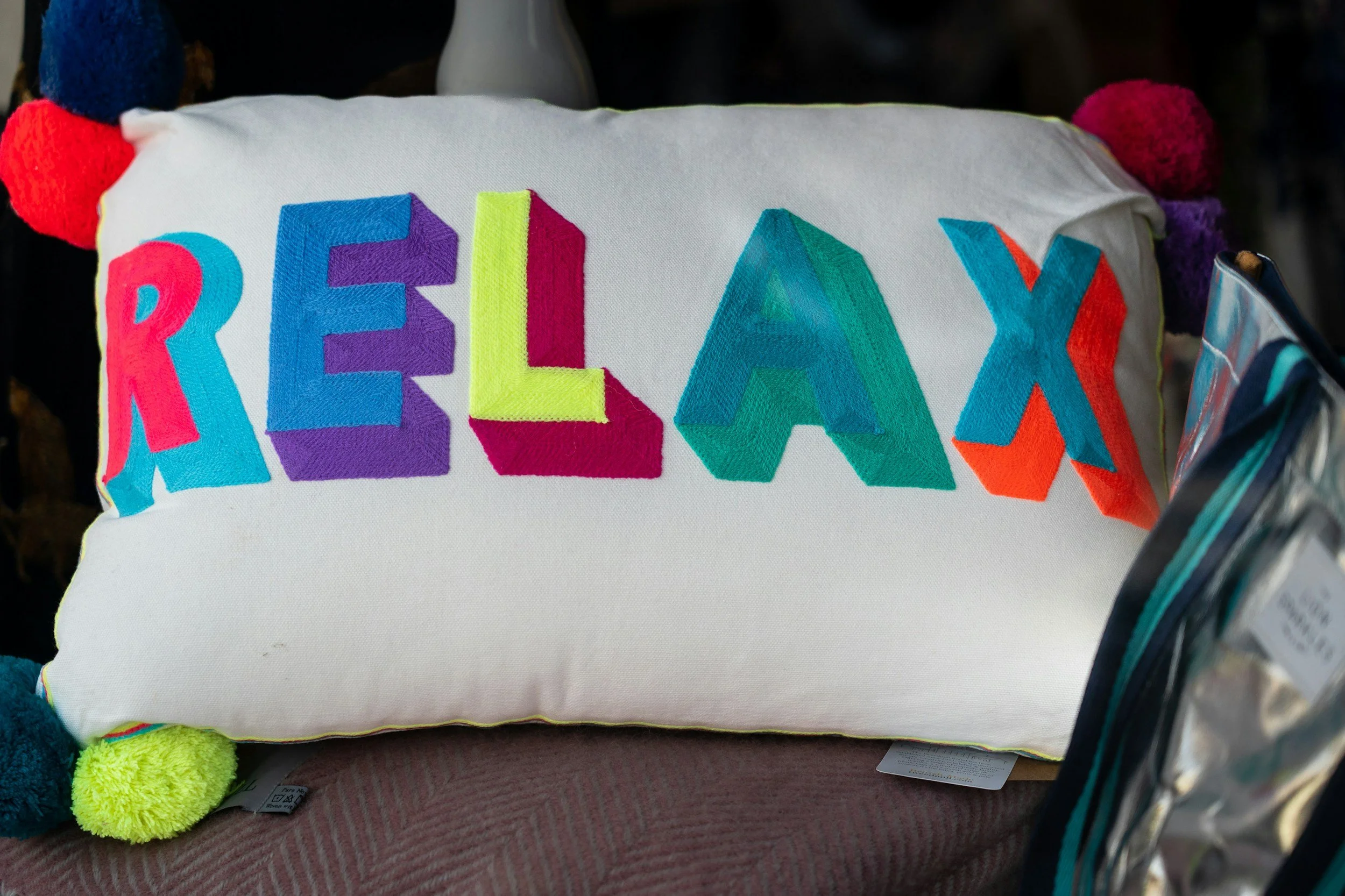A white fabric pillow with colorful embroidered letters spelling 'RELAX' and pom-pom accents on the edges.